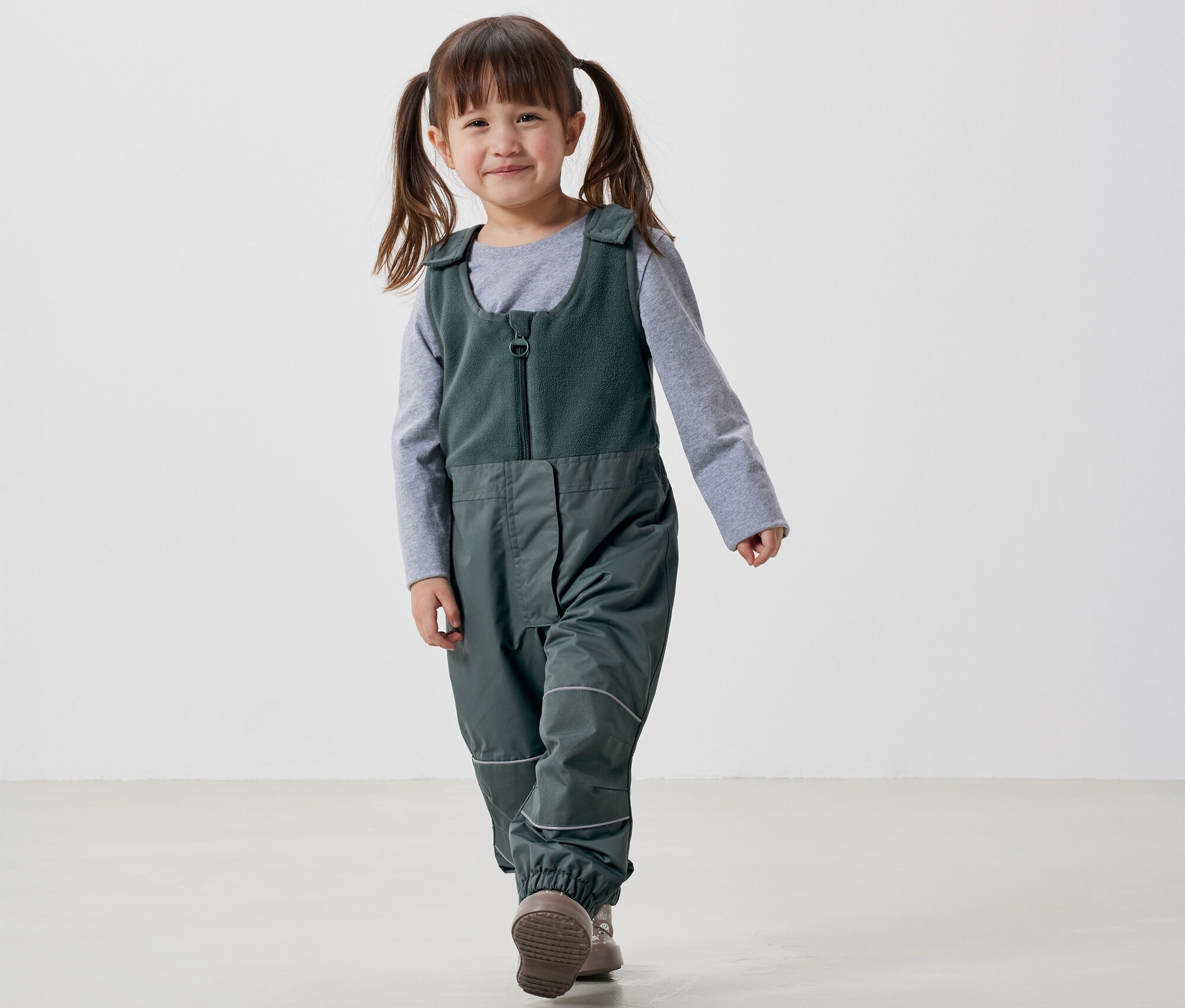 Uśmiechnięta dziewczynka z warkoczami idzie w zielonym kombinezonie i szarej koszulce na białym tle.