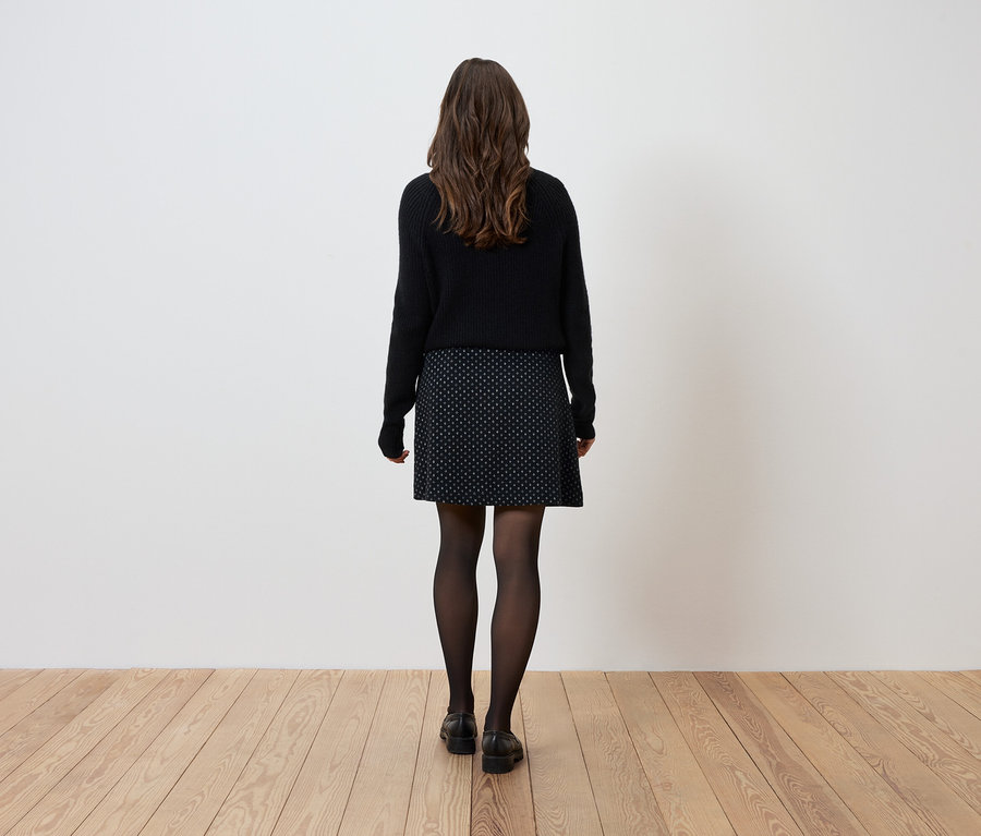 Kobieta stoi tyłem do aparatu, ubrana w czarny sweter, wzorzystą spódnicę i czarne rajstopy.