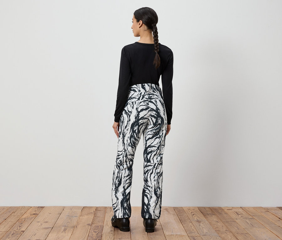 Kobieta tyłem do kamery ma na sobie czarną bluzkę z długim rękawem i biało-czarne wzorzyste spodnie.
