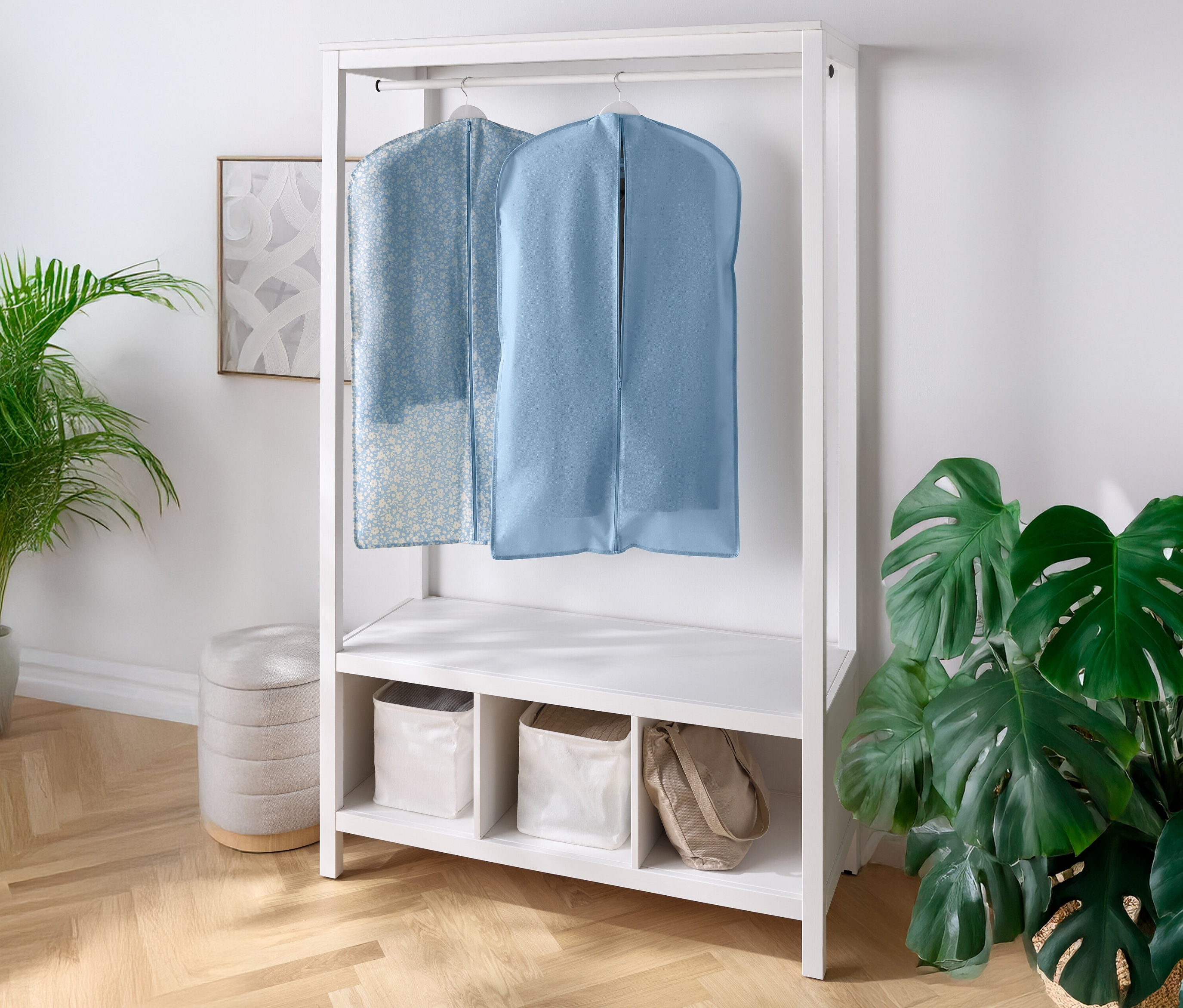 Dwa pokrowce na ubrania wiszą na białej garderobie z półkami i przegródkami pod spodem. Po lewej i prawej stronie stoi roślina doniczkowa.