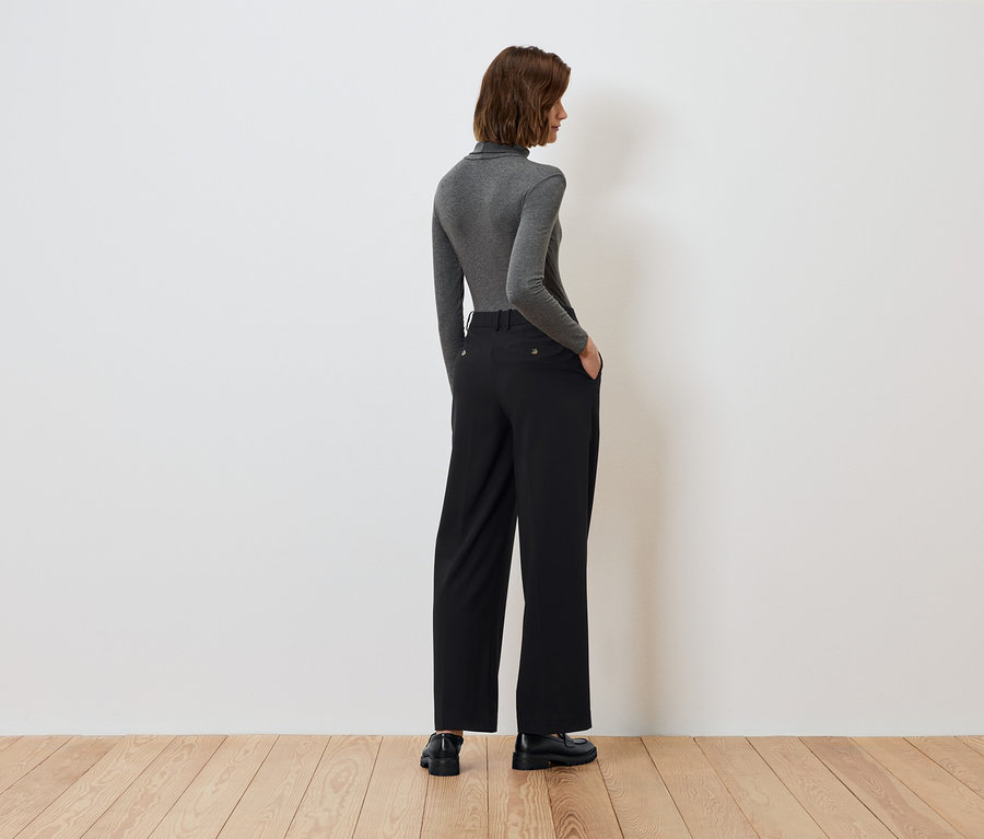 Widok z tyłu kobiety z rękami w kieszeniach czarnych spodni i szarym golfem.