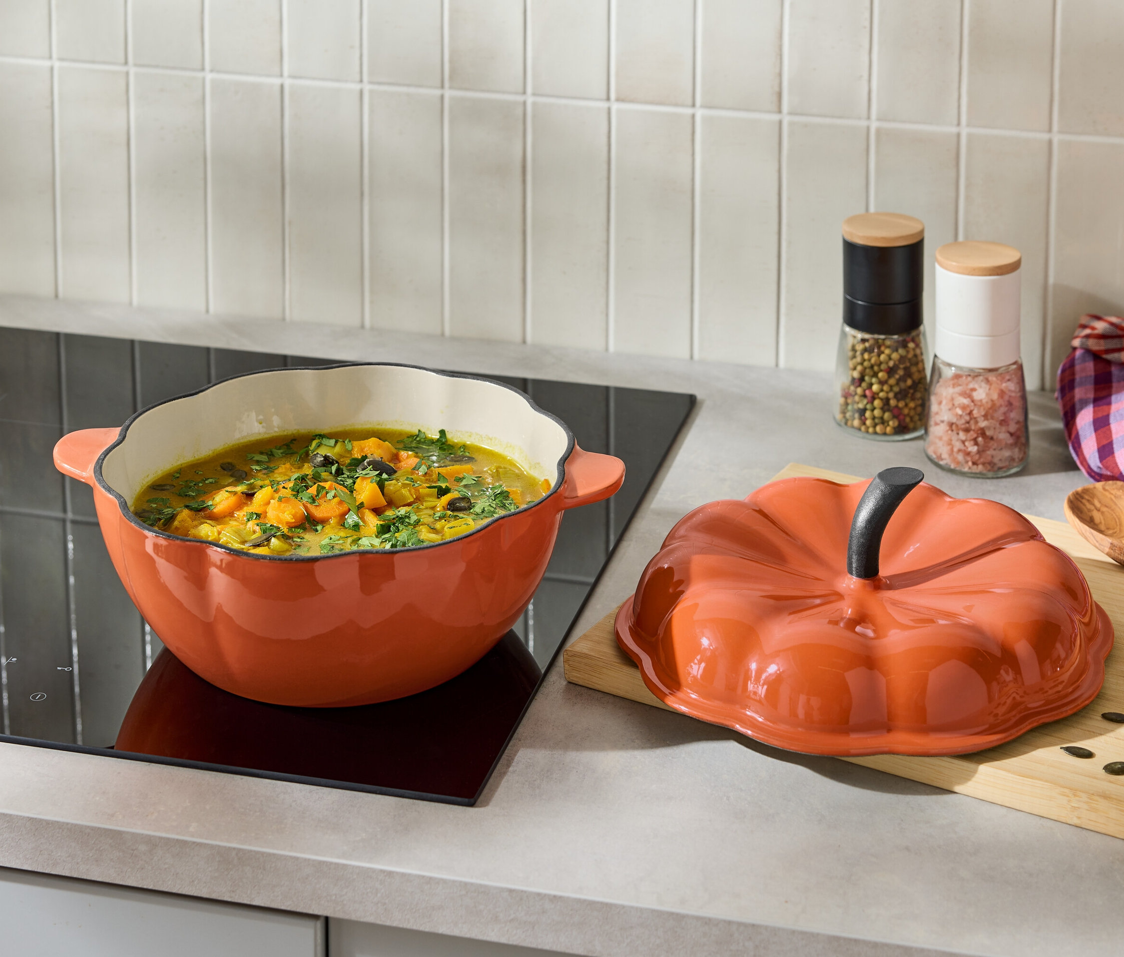 Pomarańczowy garnek z zupą na płycie indukcyjnej obok pomarańczowej pokrywki w kształcie dyni.