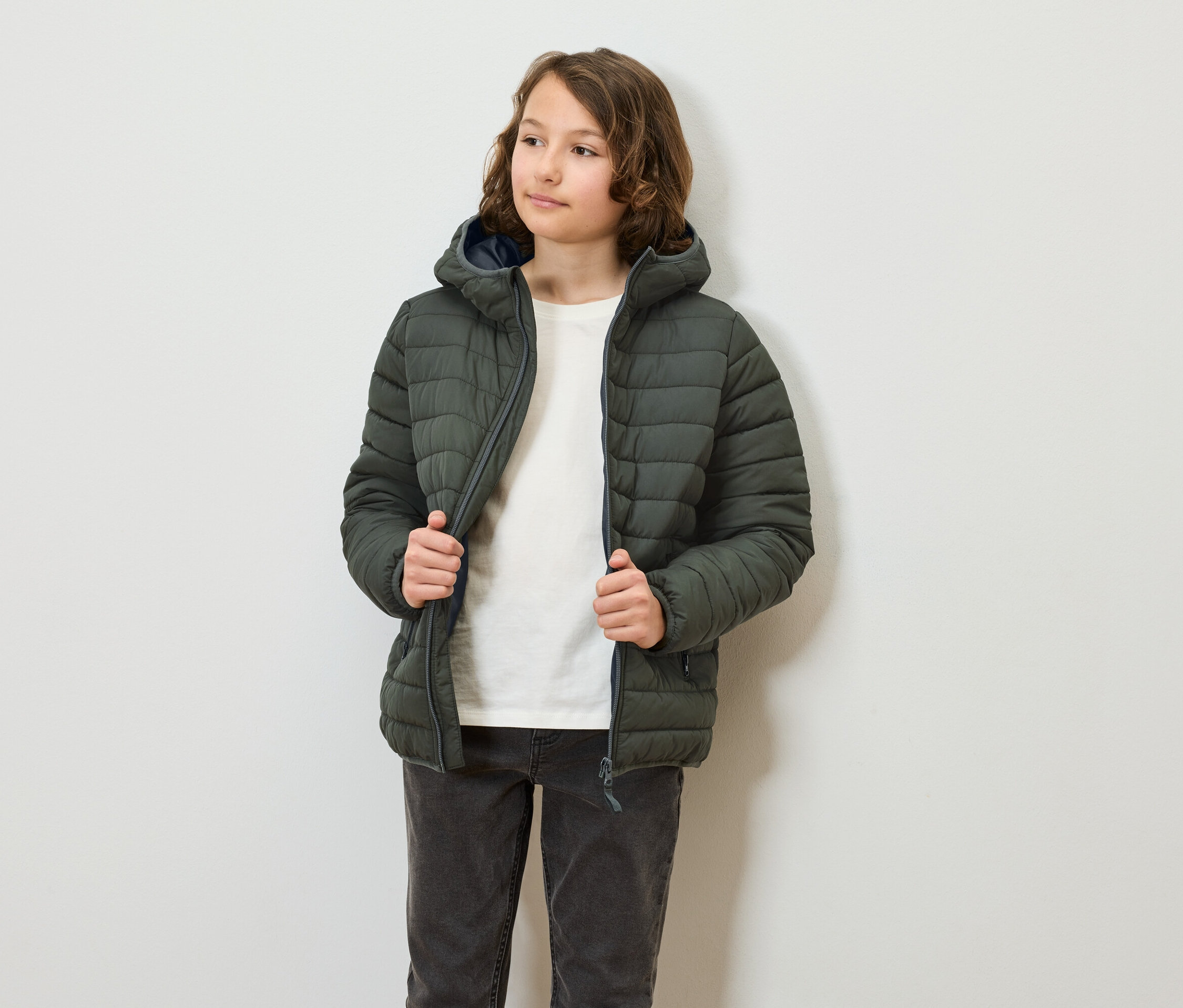 Nastolatek ma na sobie i zapina oliwkową kurtkę puchową z kapturem. Osoba ma na sobie dżinsy i t-shirt.