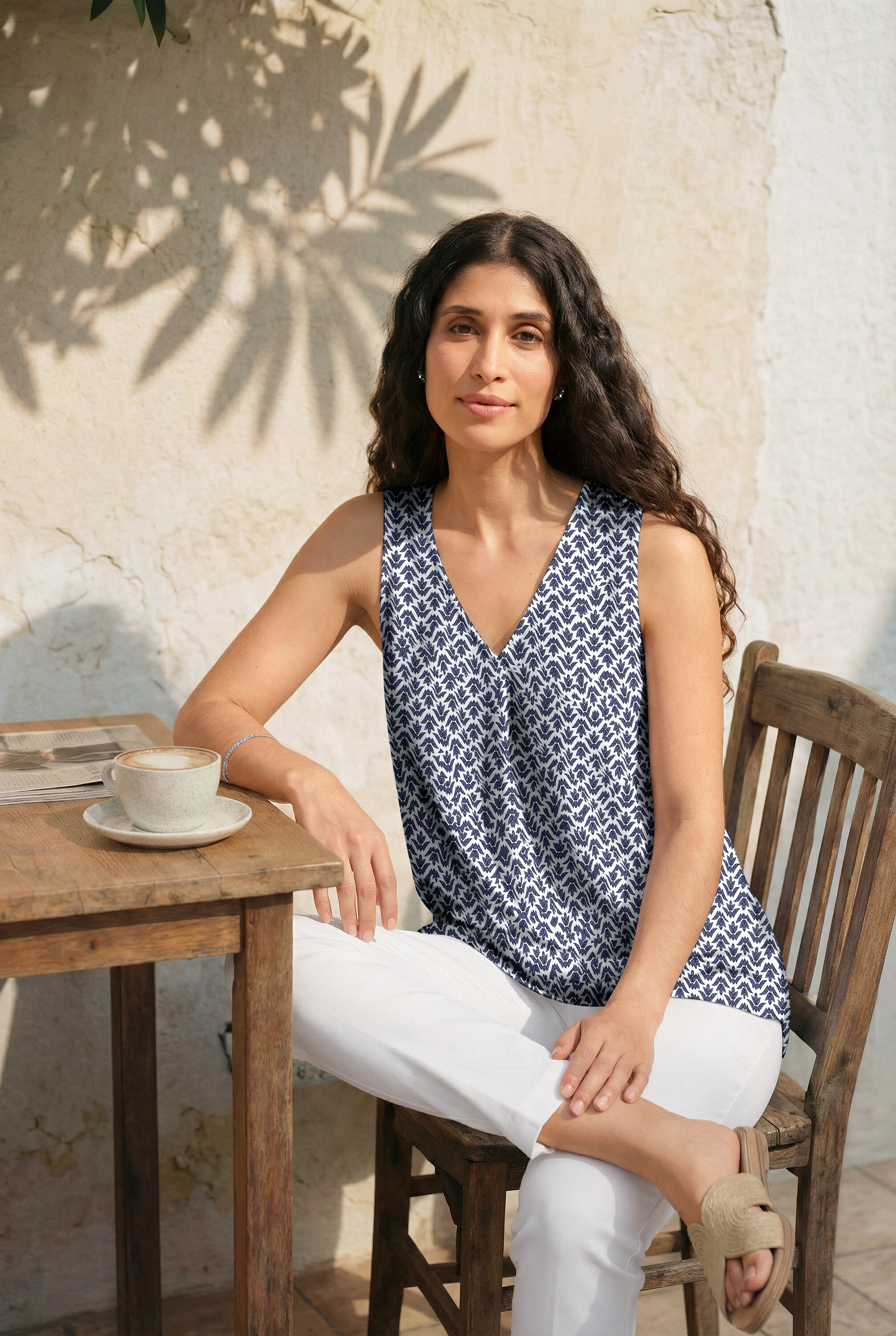 Kobieta siedzi przy stole w kolczykach i z bransoletką tenisową. Ma na sobie top z nadrukiem, białe spodnie 7/8 i klapki.