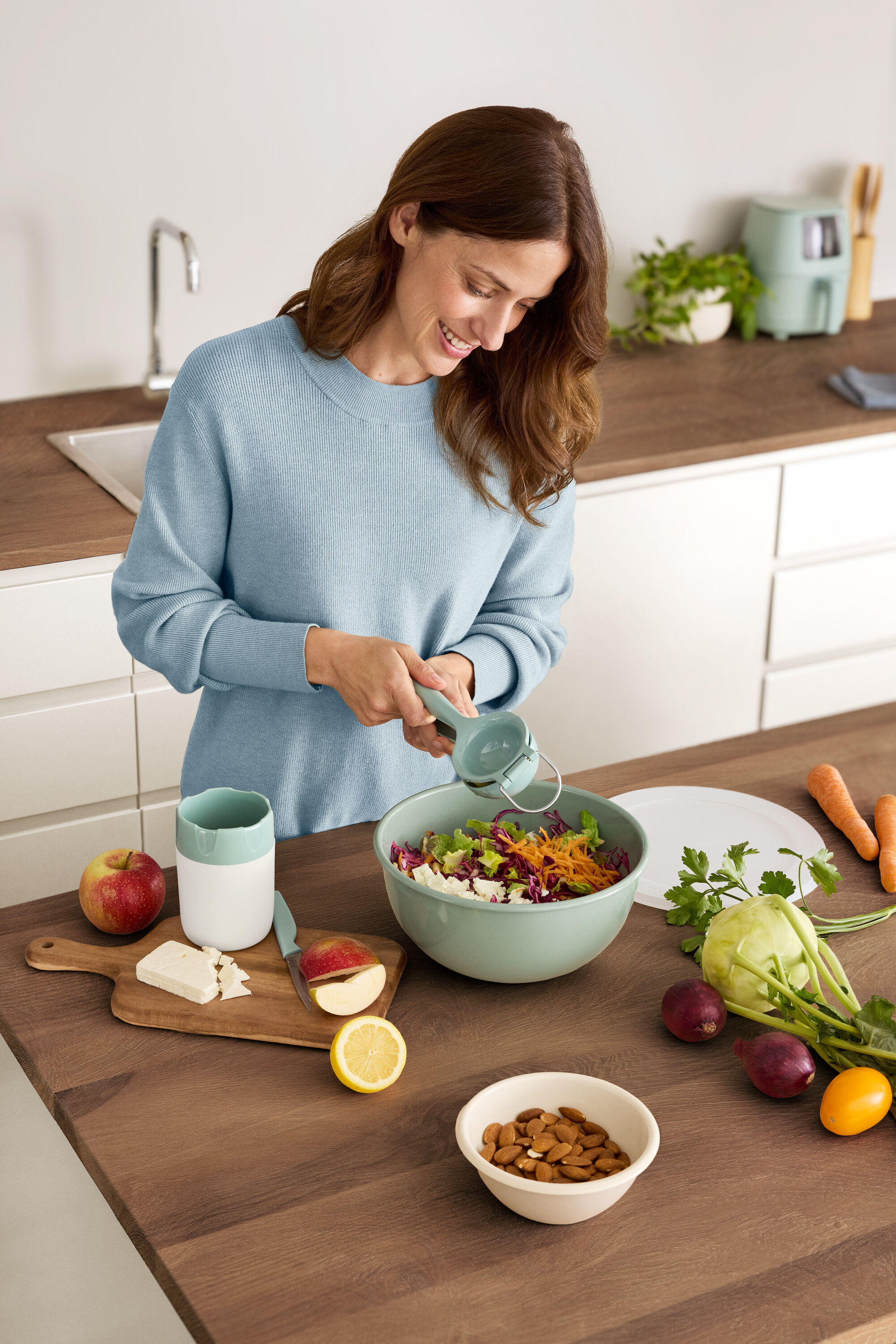 Kobieta wyciska cytrynę do miski sałatki. Na stole leżą jabłko, ser, migdały i warzywa.