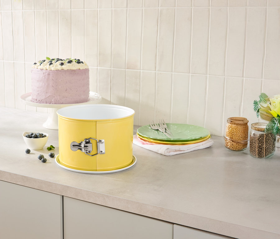 Na blacie kuchennym stoi żółta forma do pieczenia, ciasto na paterze, talerze i słoiki z zapasami.