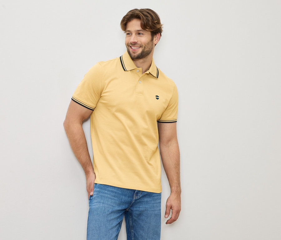 Mężczyzna w żółtej koszulce polo i dżinsach stoi na tle jasnej ściany.