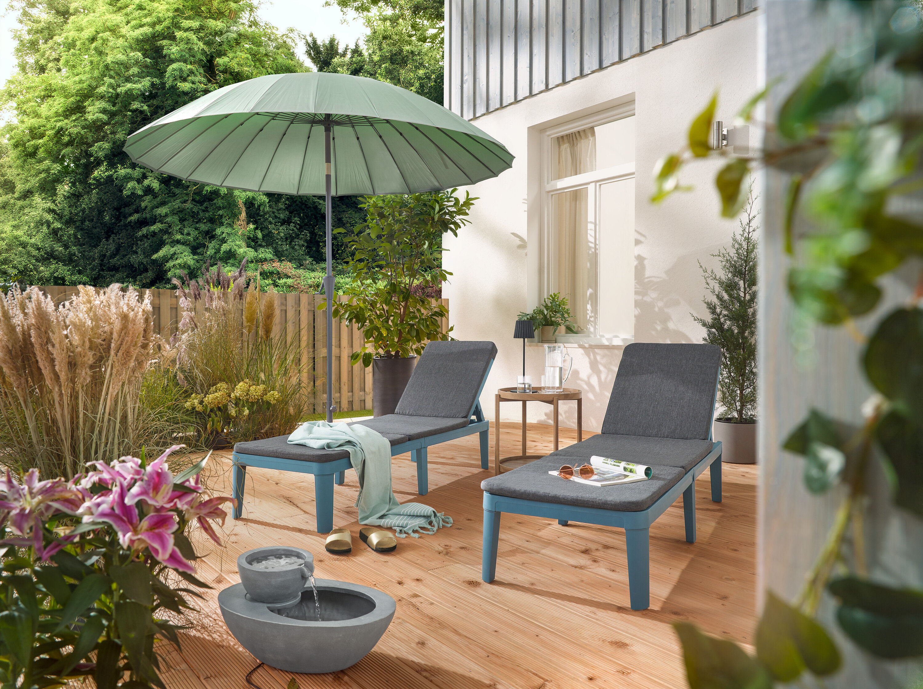 Na tarasie stoją dwa leżaki pod jasnoszarym parasolem Leco Shanghai i obok solarnej fontanny ogrodowej.