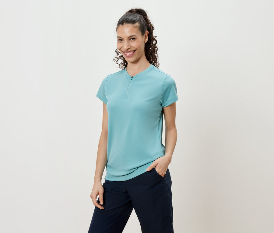 Kobieta z kręconymi włosami związanymi w kucyk, ubrana w turkusową koszulkę i granatowe spodnie.