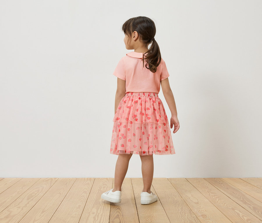 Dziewczynka stoi tyłem do kamery, ma na sobie różowy T-shirt, różową tiulową spódniczkę w truskawki i białe tenisówki.