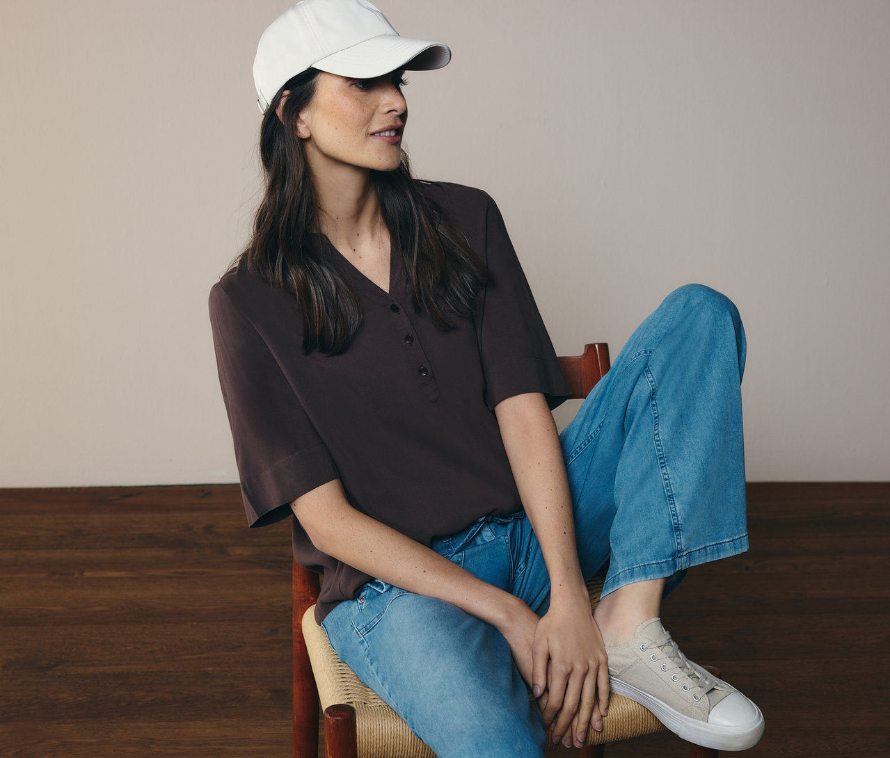 Kobieta siedzi na krześle, ma na sobie dżinsy, czapkę i brązową bluzkę tunikową CECIL.