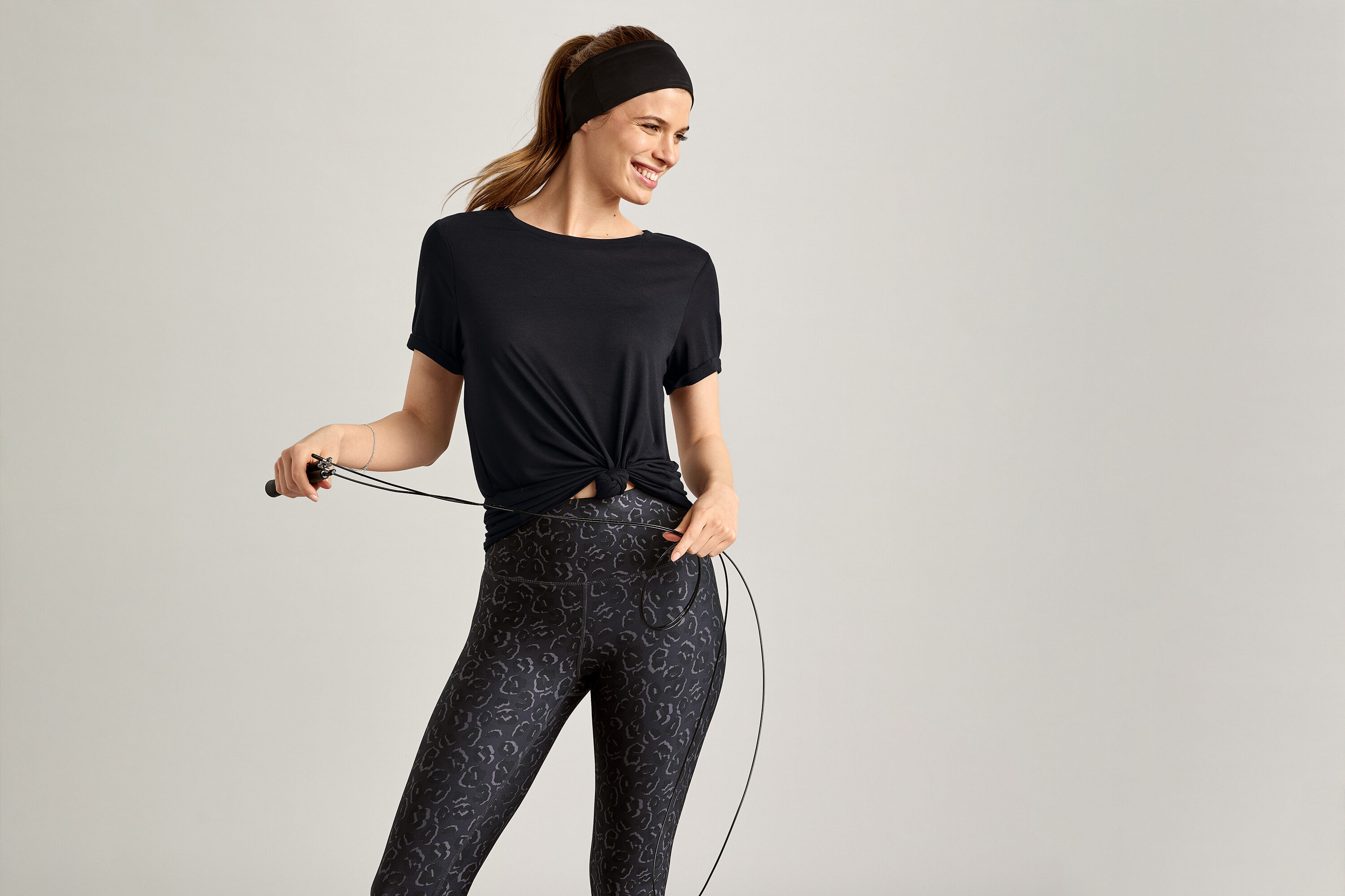 Uśmiechnięta kobieta w czarnej koszulce sportowej trzyma skakankę na szarym tle.