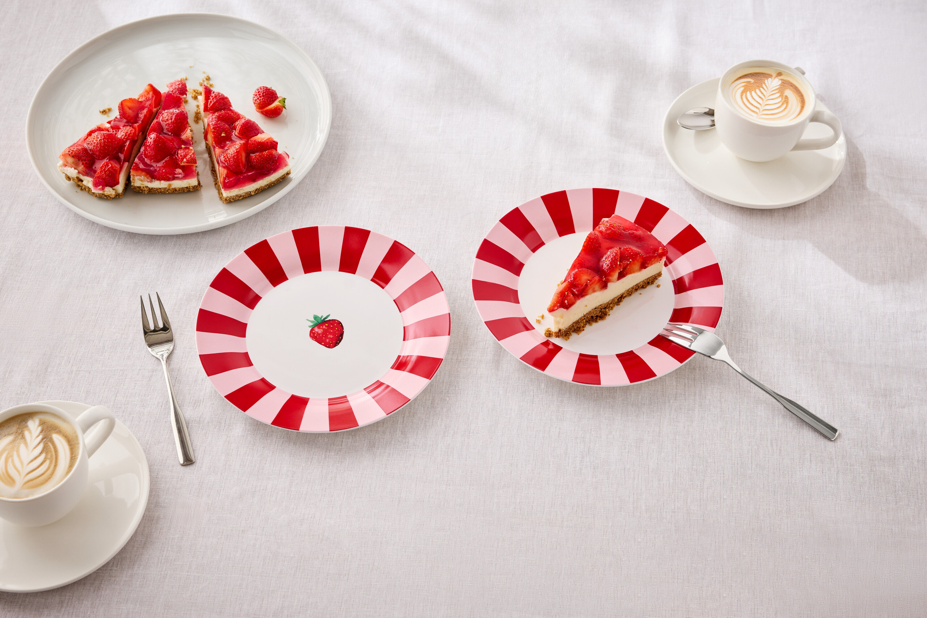 Na stole leżą talerze z ciastem truskawkowym, widelce i filiżanki z latte art.