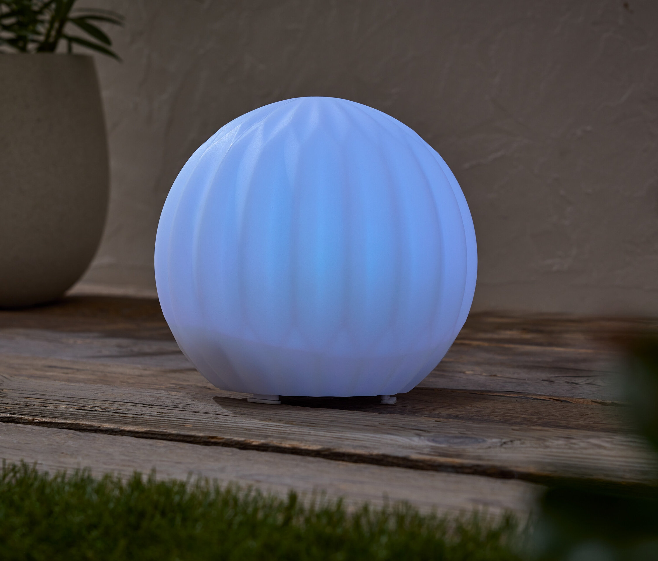 Świecąca, kulista lampa stoi na drewnianym stole obok doniczki z rośliną.