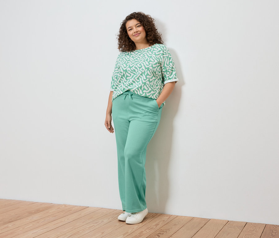 Kobieta pozuje przy ścianie, ubrana w jasnozieloną bluzkę we wzory, jasnozielone spodnie i białe tenisówki.