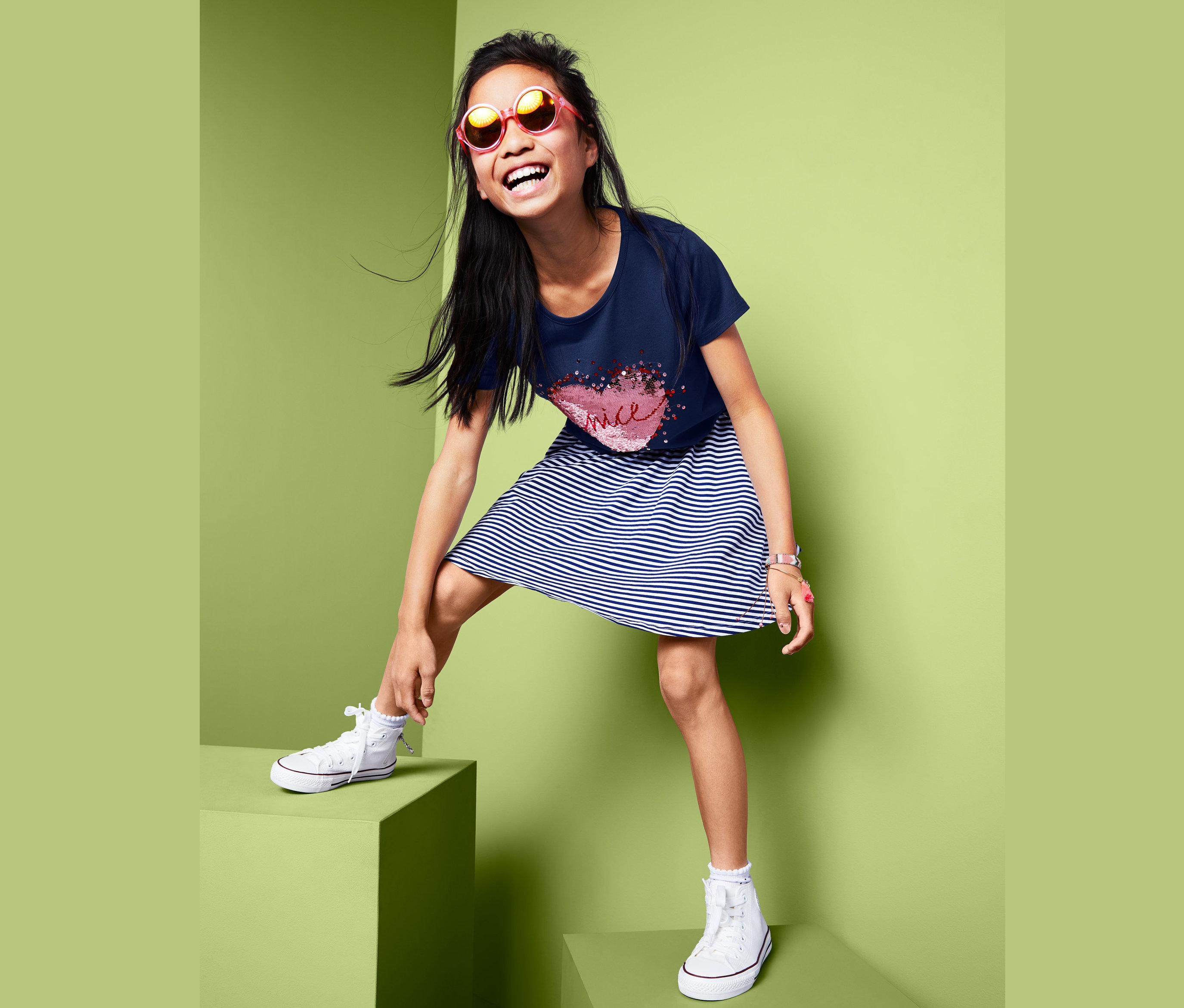 Uśmiechnięta dziewczyna w koszulce dziewczęcej z krótkim rękawem z dwustronnymi cekinami, w pasiastą sukienkę i trampki stoi na zielonych kostkach.