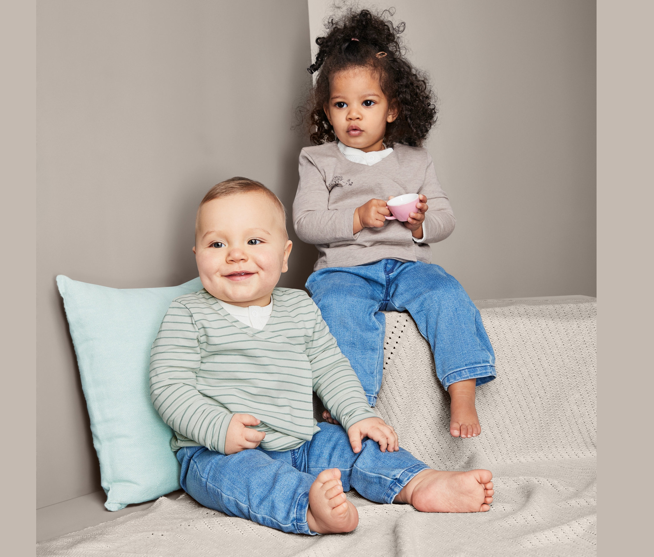 Dziecko siedzi obok malucha. Maluch trzyma różowy kubek. Oboje mają na sobie dżinsy i dżersejowe koszulki z krojem kopertowym, 2 sztuki.