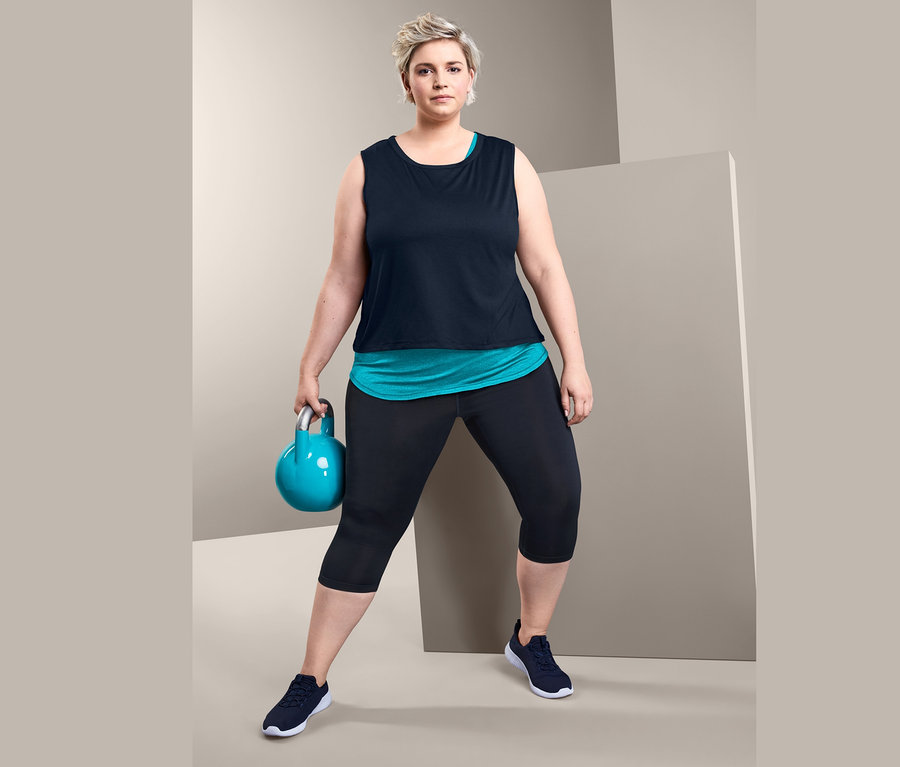 Kobieta trzyma kettlebell i ma na sobie top sportowy 2 w 1 oraz legginsy sportowe o długości 3/4.