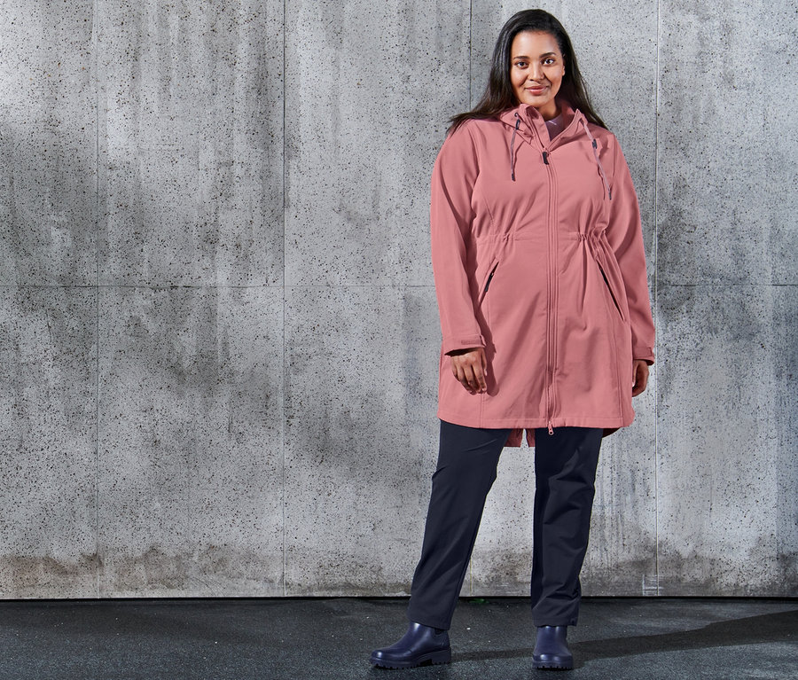 Kobieta pozuje na tle betonowej ściany, ubrana w płaszcz softshell i granatowe spodnie dresowe softshell.