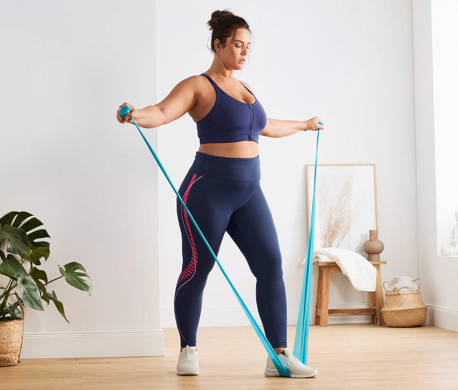 Kobieta ćwiczy z niebieską gumą oporową. Ma na sobie legginsy sportowe i biustonosz sportowy z zamkiem błyskawicznym z przodu.