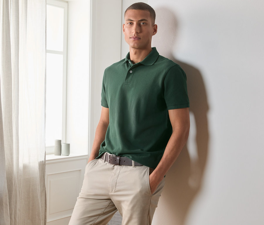 Mężczyzna pozuje oparty o ścianę, ubrany w ciemnozieloną koszulkę polo o splocie piqué.