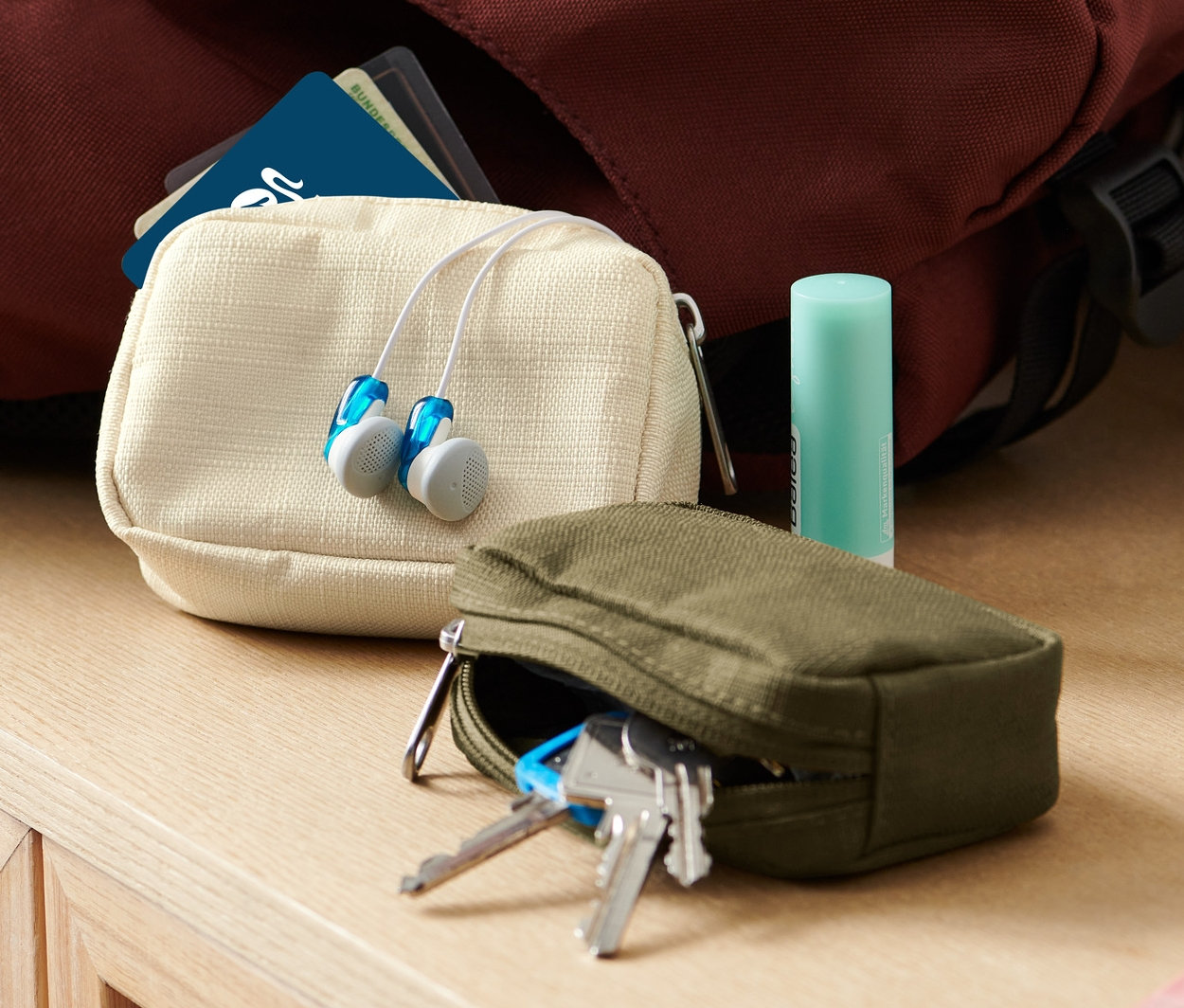 Dwa mini organizery podróżne, słuchawki i balsam do ust leżą na komodzie obok plecaka i wazonu.