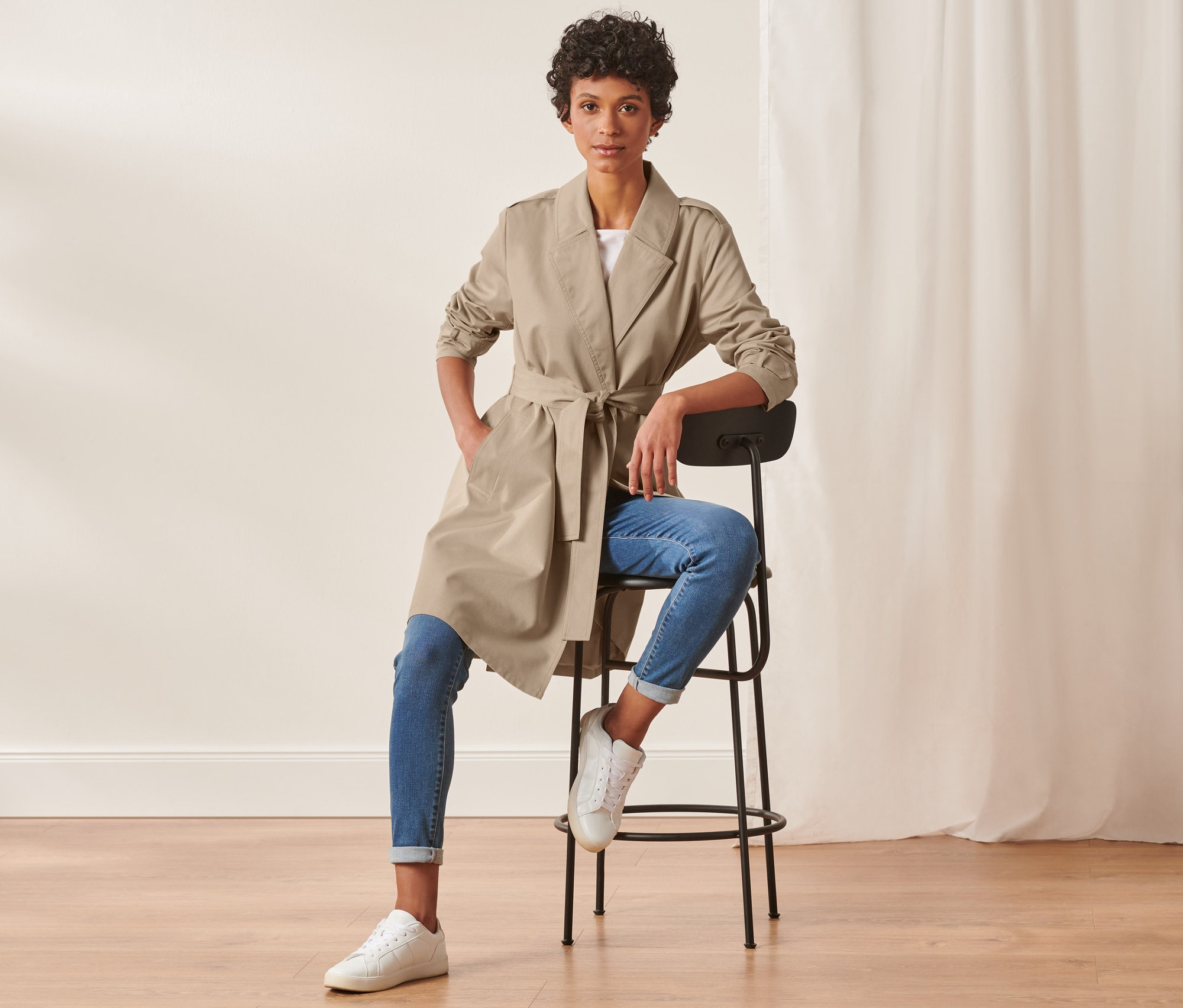 Kobieta siedzi na krześle w trenczu, dżinsach i tenisówkach.