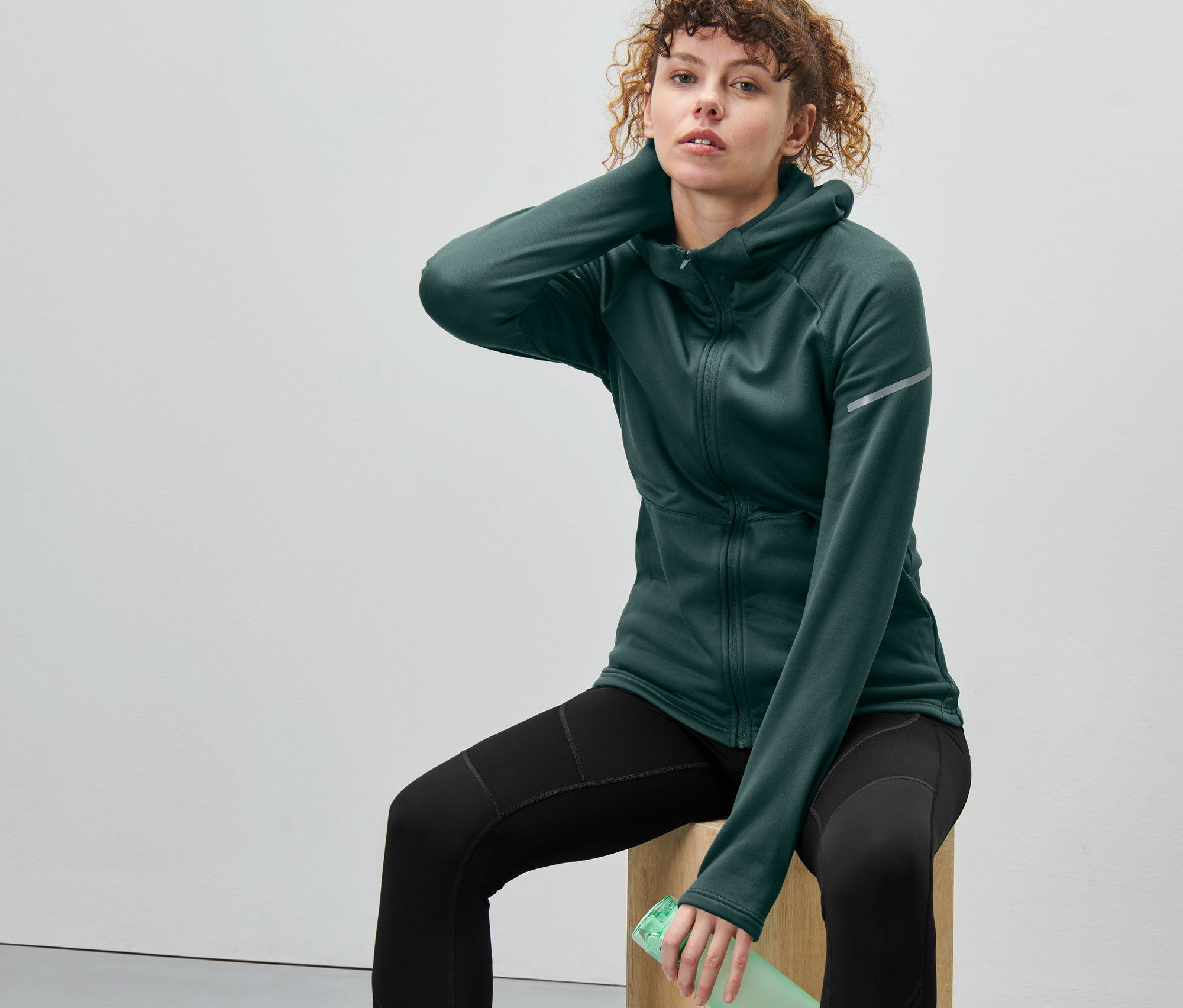 Kobieta siedzi na drewnianym bloku, ubrana w ciemnozieloną kurtkę termoaktywną.