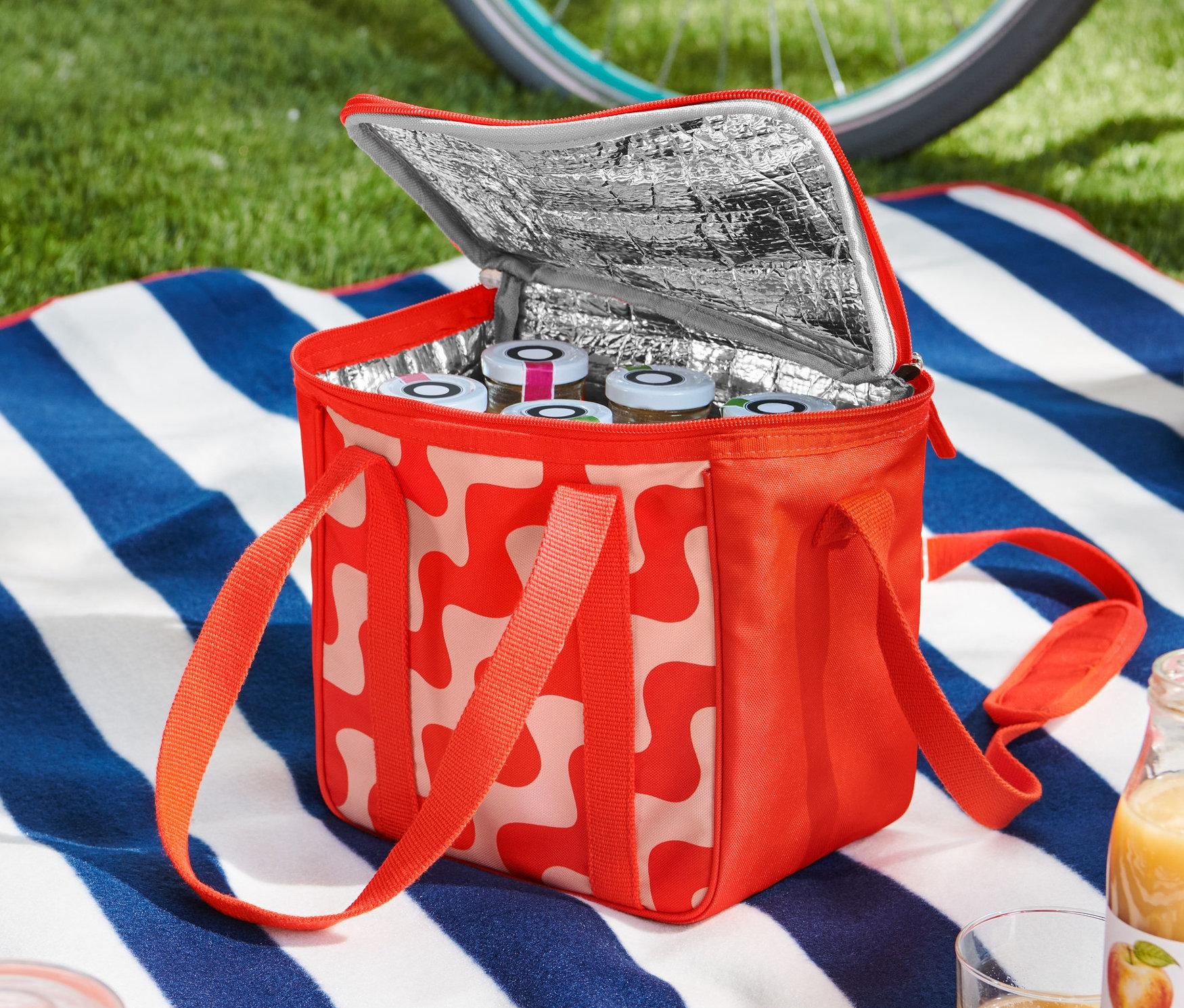 Otwarta mała torba chłodząca w czerwono-biały wzór stoi na niebiesko-białym kocu w paski, wypełniona słoikami. W tle widać rower i kask.