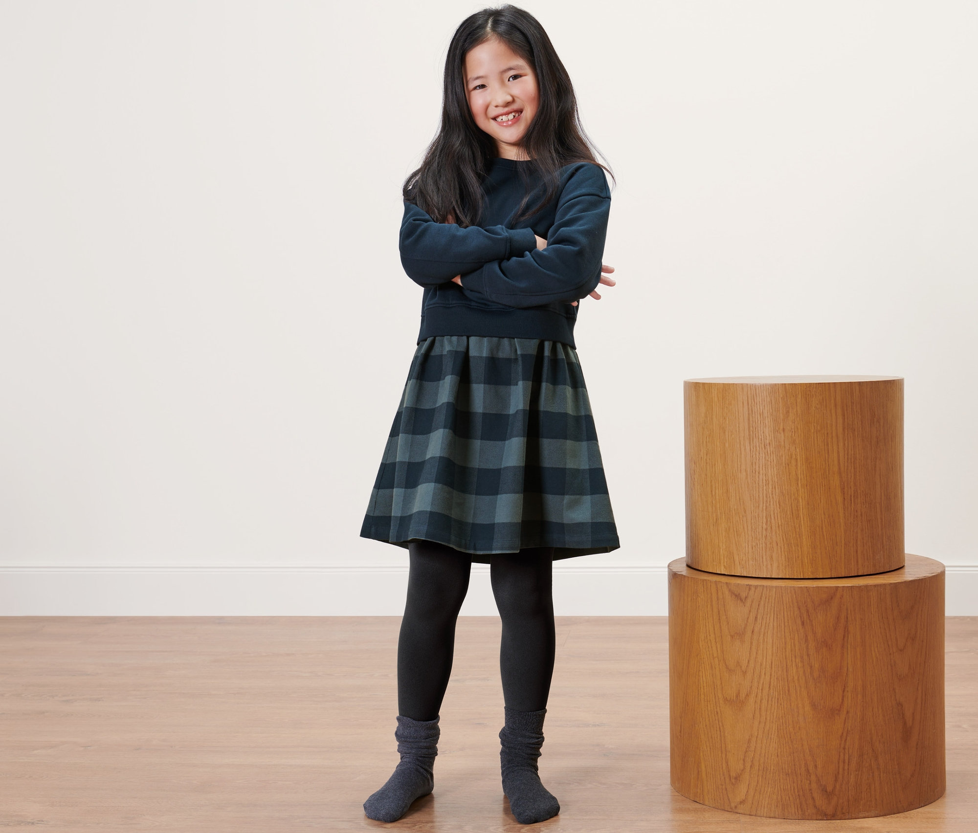 Uśmiechnięta dziewczyna pozuje ze skrzyżowanymi ramionami, ubrana w legginsy termiczne dla dzieci, sweter i spódnicę w kratę.