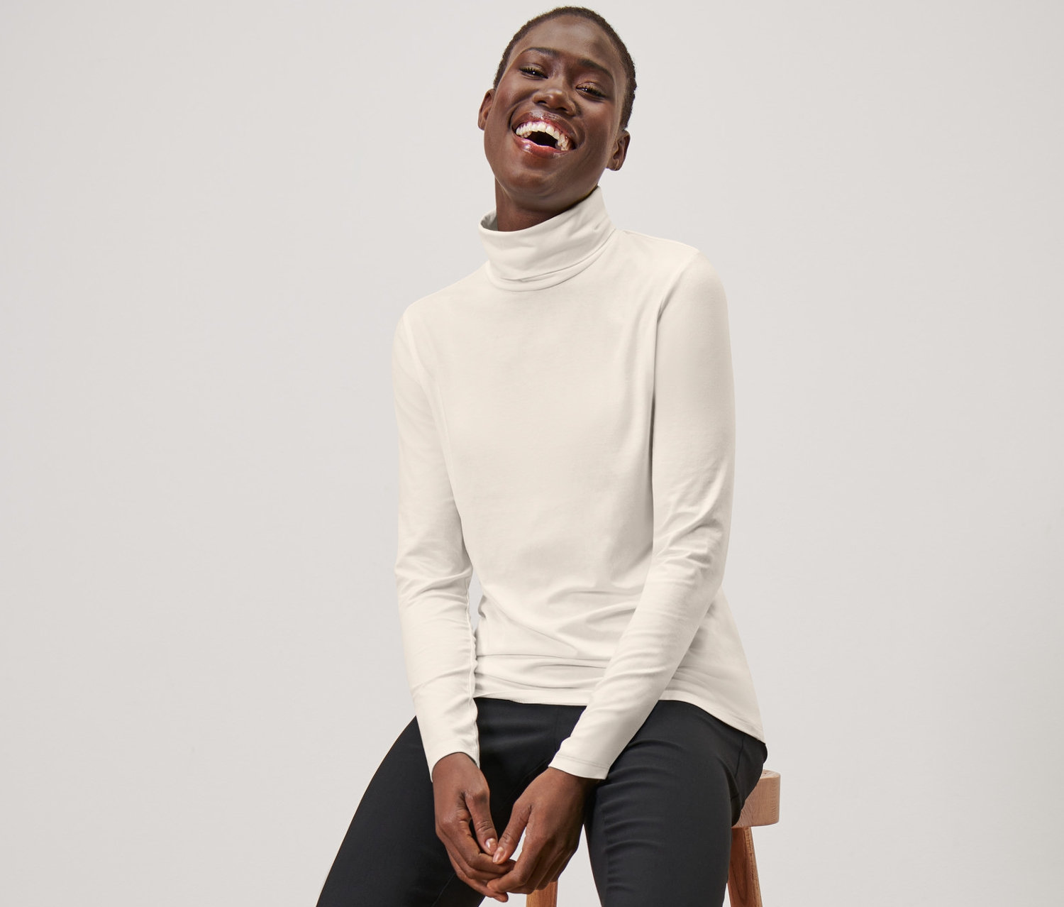 Kobieta siedzi na stołku, ubrana w kremowo-białą bluzkę z kołnierzem golfowym, czarne spodnie i czarne mokasyny.