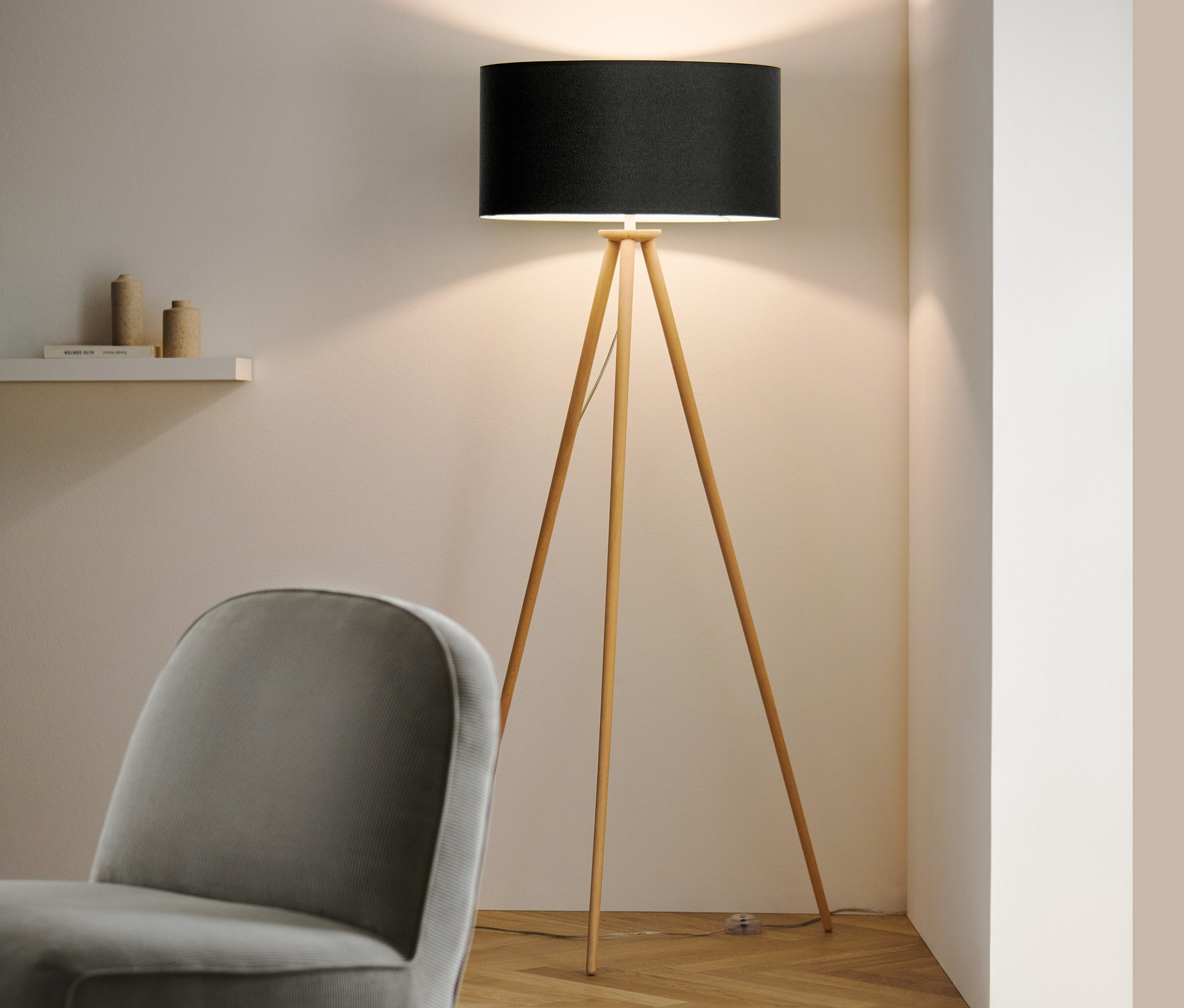 W pokoju stoi drewniana lampa stojąca „trójnóg” z czarnym abażurem i szary fotel.