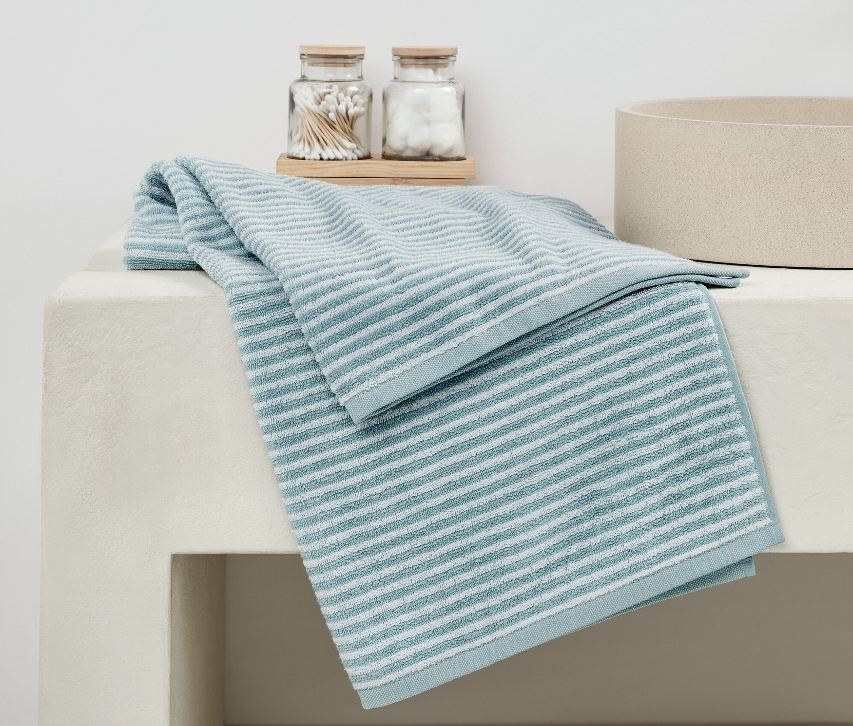 Wysokiej jakości niebieski ręcznik kąpielowy leży złożony na białej umywalce obok szklanek z wacikami i kulkami.