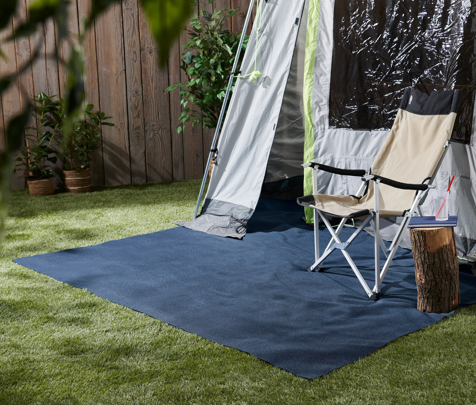 Niebieski dywan do namiotu o wymiarach 220 x 350 cm leży na trawie przed namiotem. Obok stoi krzesło kempingowe i pień drzewa służący jako stolik.