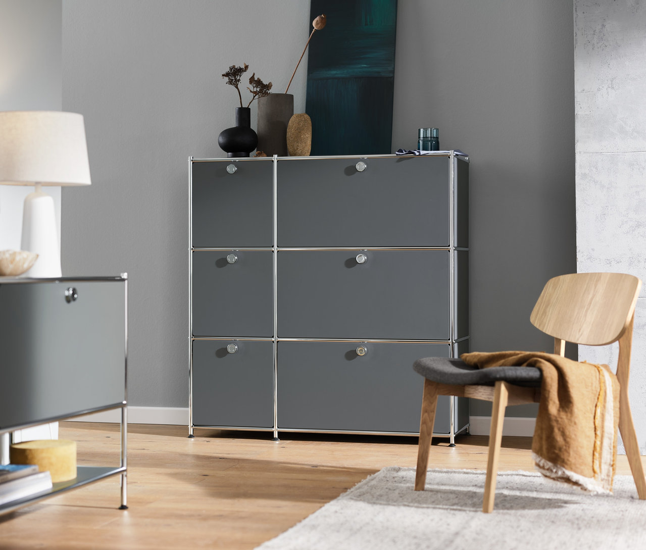 Wnętrze z szarą komodą »CN3« z 6 klapami, szarą szafką typu sideboard »CN3« z 1 klapą, białą lampą stołową i krzesłem.