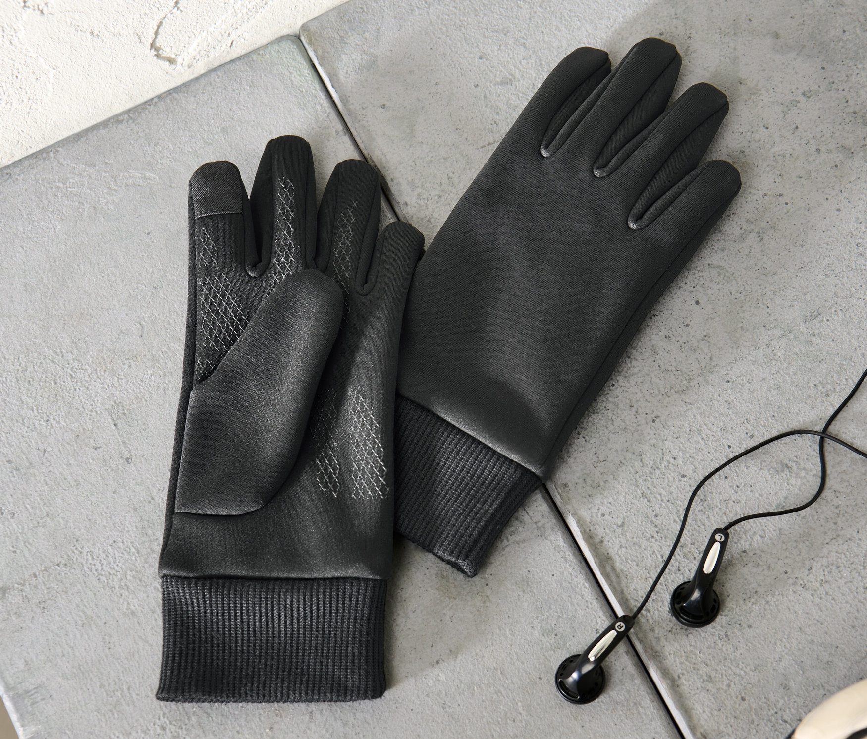 Dwie dziecięce rękawiczki termoaktywne chroniące przed wiatrem leżą na szarej powierzchni obok słuchawek i części piłki nożnej.