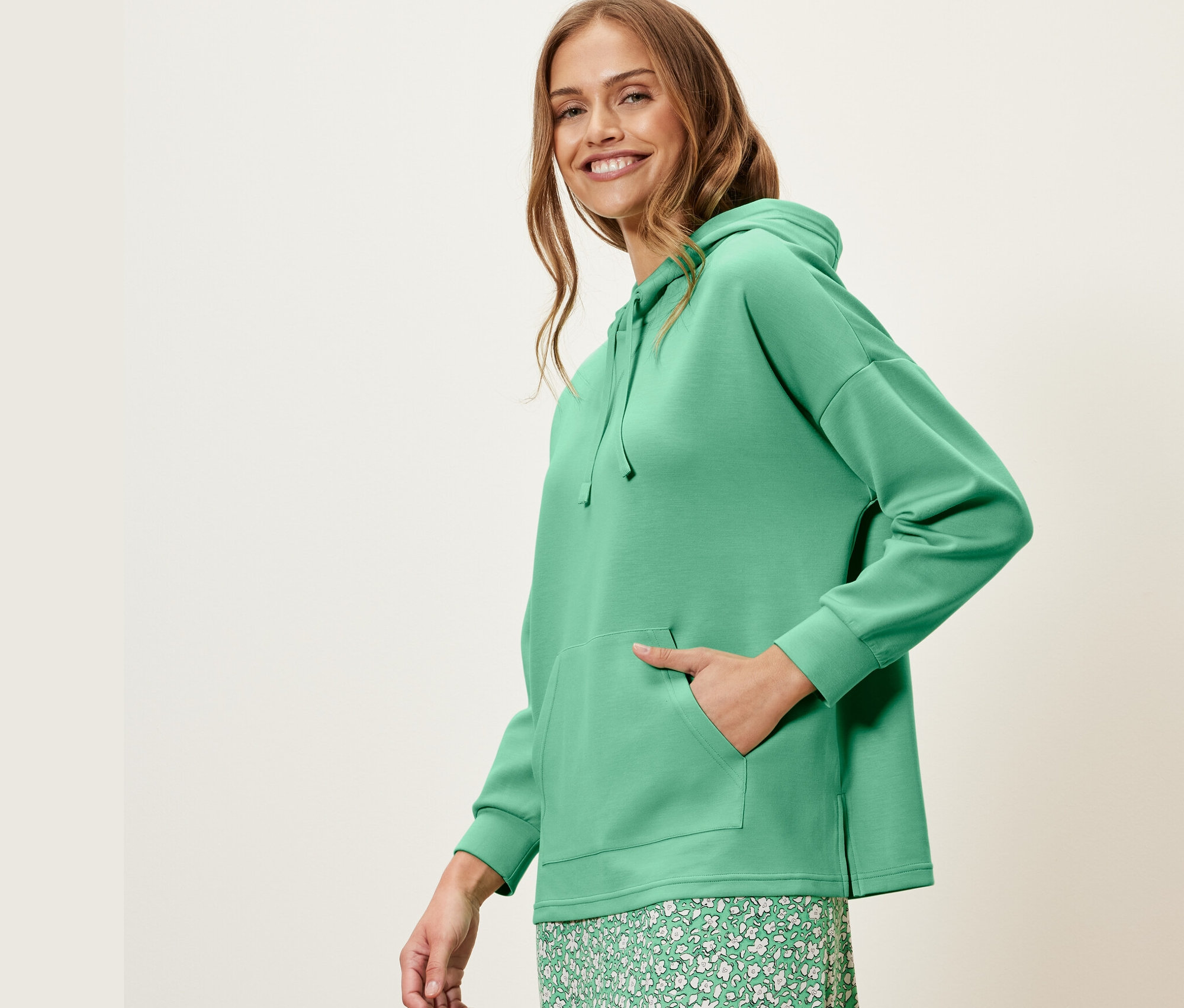 Kobieta pozuje w zielonej bluzie z kapturem z dodatkiem modalu i zielonej kwiecistej spódnicy.