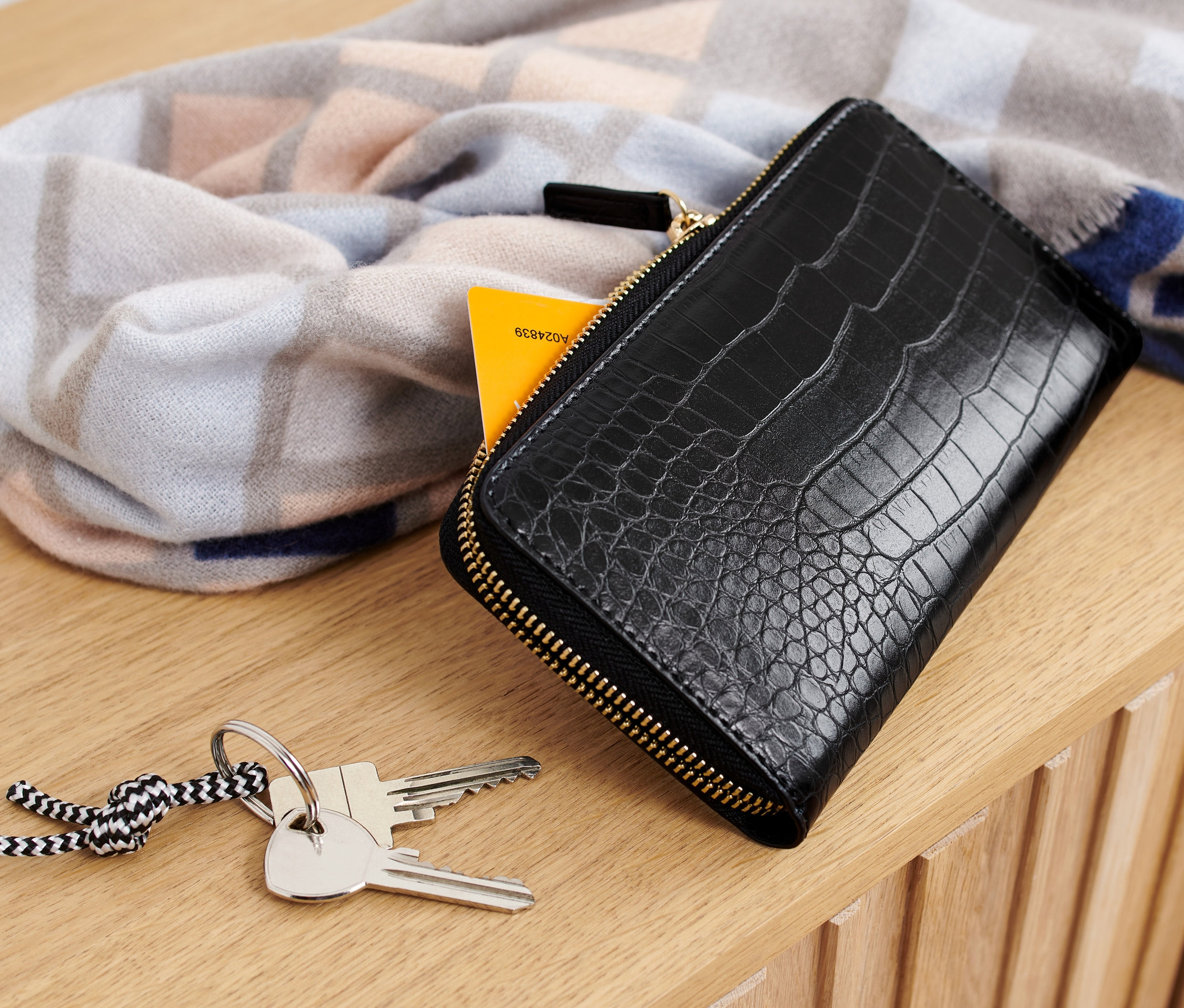 Czarny portfel leży na drewnianej powierzchni obok kluczy i wzorzystego szala.