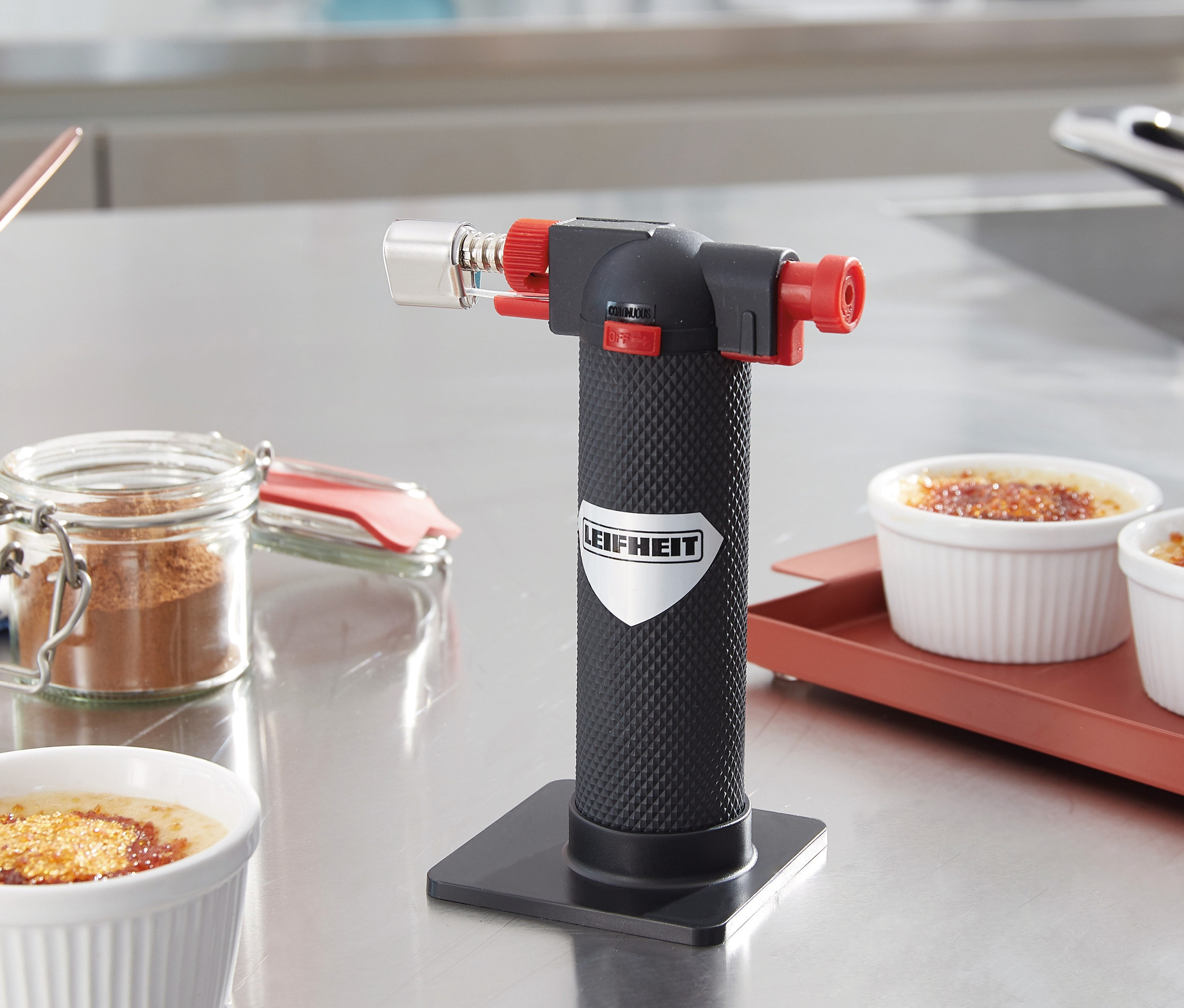 Na stole stoi palnik Gourmet i palnik kuchenny obok misek z crème brûlée.