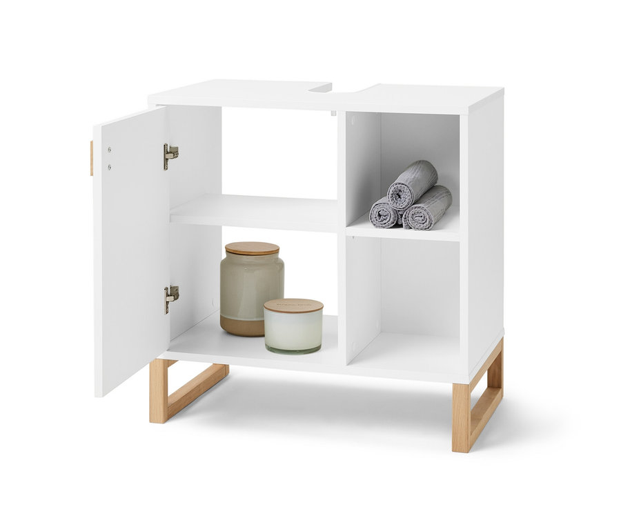 Otwarta, biała szafka pod umywalkę z drzwiczkami i boczną przegródką. W bocznej przegródce zwinięte szare ręczniki, wewnątrz szafki świece i puszka.