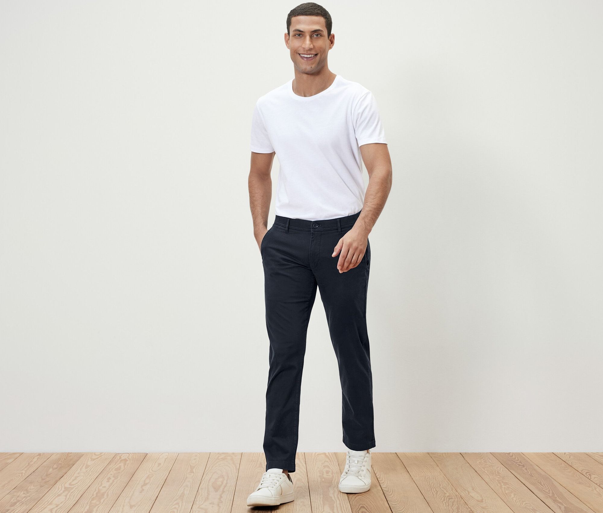 Mężczyzna pozuje do kamery w białym T-shircie i granatowych spodniach chinosach.