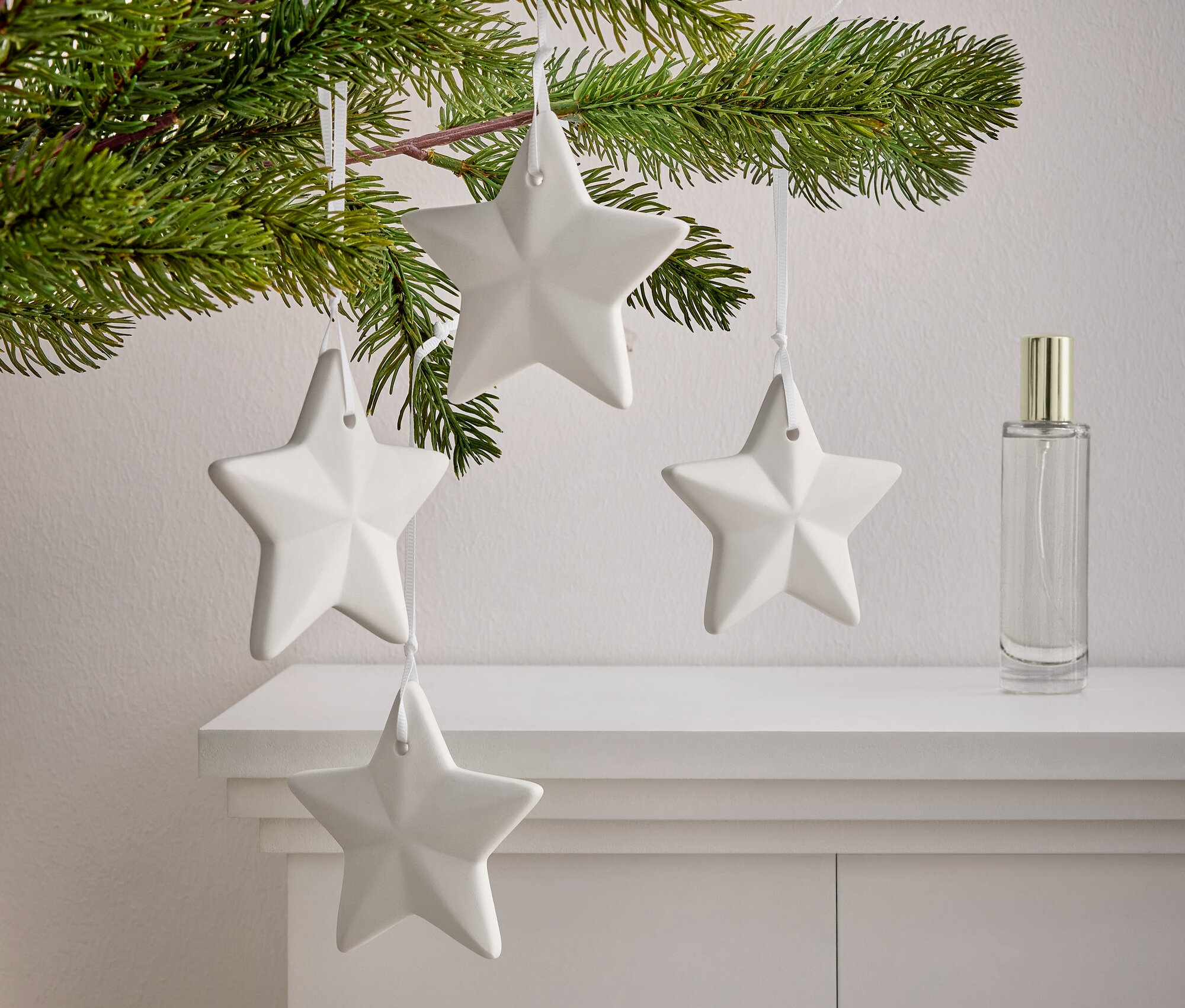 Gałązka z igłami, na której wiszą cztery białe gwiazdy na białej szafce. Obok gwiazd stoi flakon perfum.