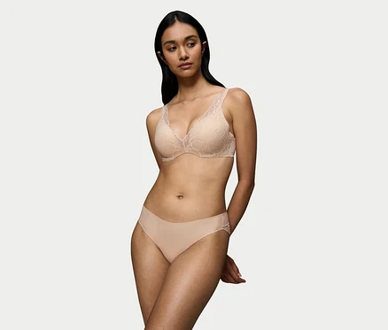 Kobieta z ciemnymi włosami nosi beżowy koronkowy biustonosz Triumph Body Makeup Illusion Lace WP z kwiatowym wzorem i pasujące beżowe figi. Jej ręce są splecione za plecami.