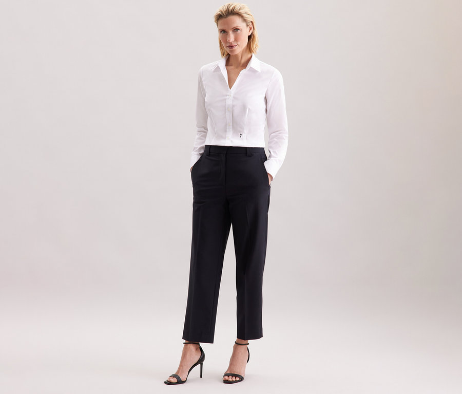 Kobieta stoi i ma na sobie białą bluzkę Seidensticker Slim Fit non iron, czarne spodnie i czarne szpilki.