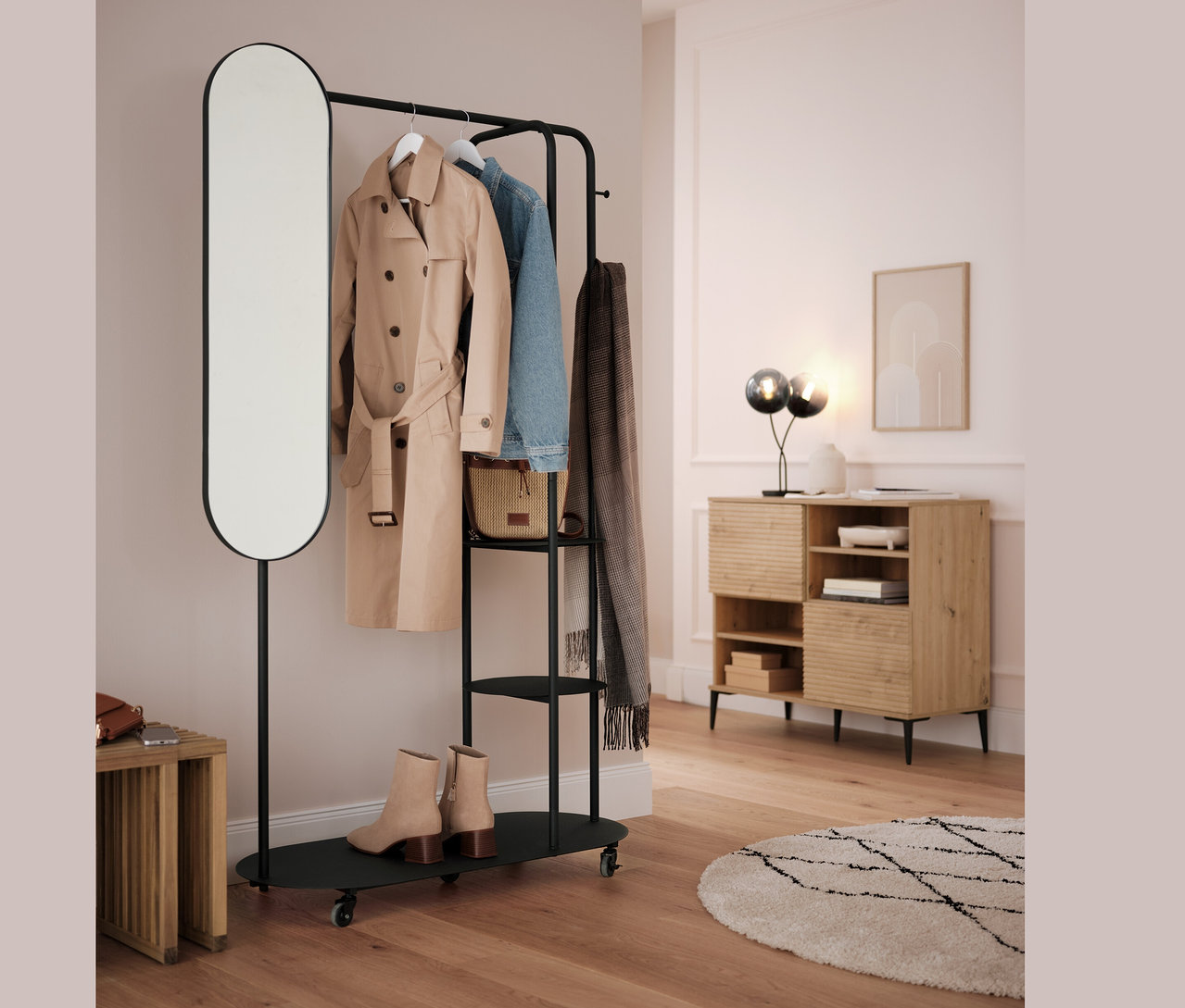 Czarny wieszak na ubrania z owalnym lustrem i rozkładanym stołkiem. Na wieszaku wisi beżowy płaszcz, niebieska kurtka i szary szal.