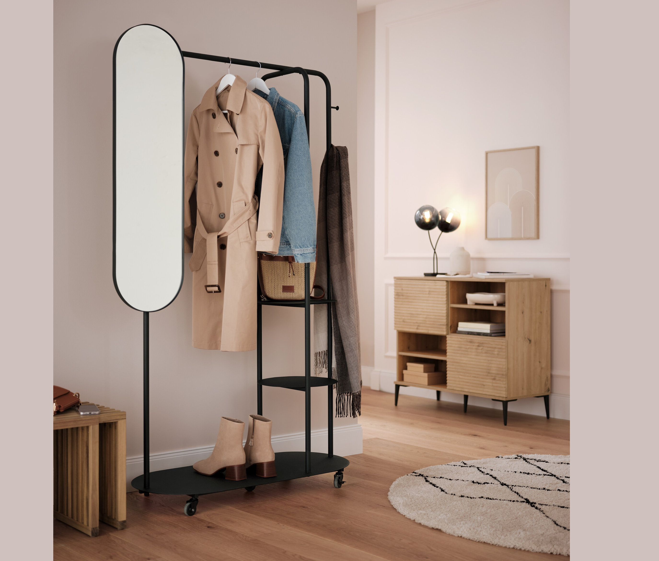 Czarny wieszak na ubrania z owalnym lustrem i rozkładanym stołkiem. Na wieszaku wisi beżowy płaszcz, niebieska kurtka i szary szal.