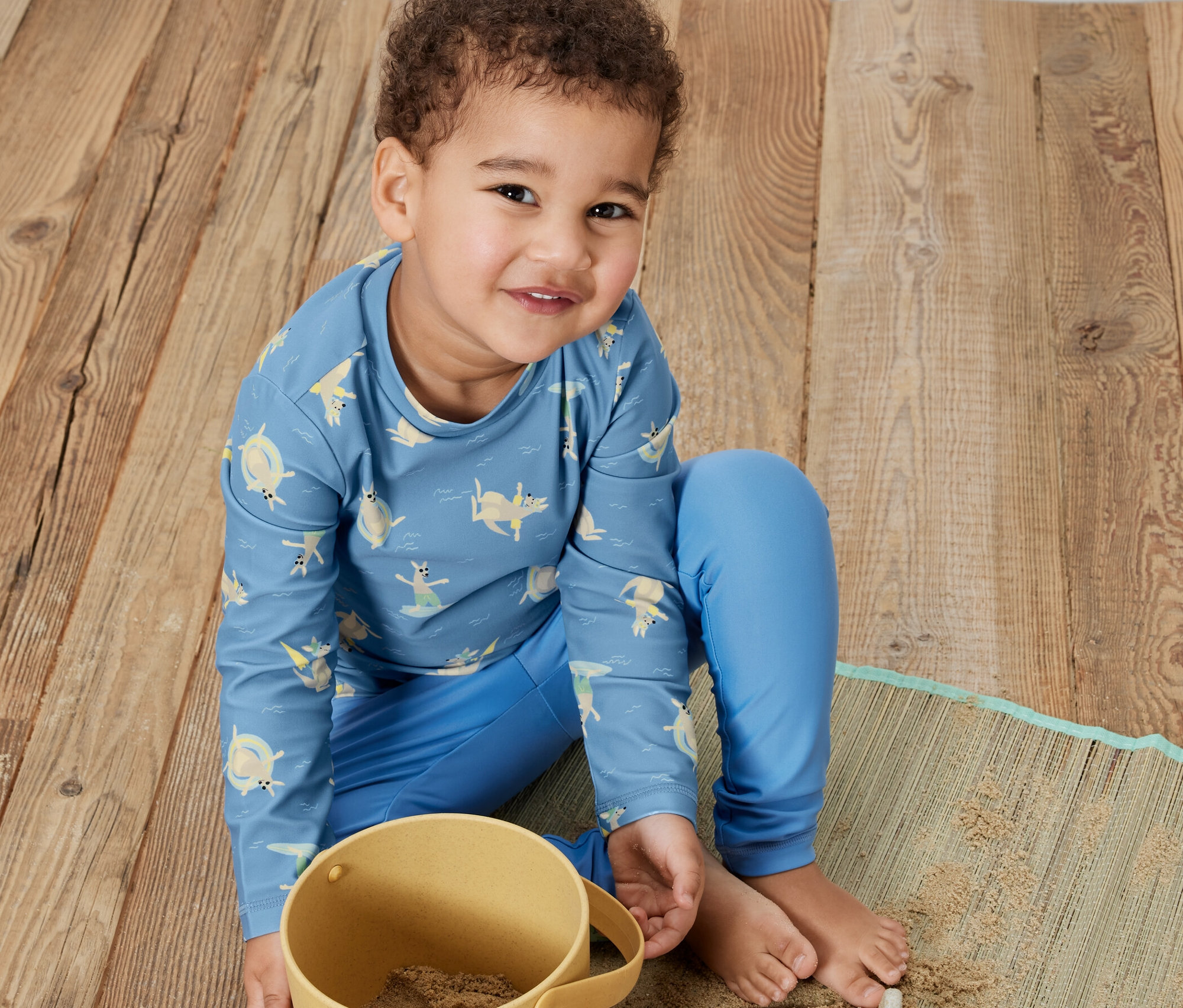 Chłopiec siedzi na drewnianej podłodze i bawi się piaskiem w żółtym wiaderku. Ma na sobie niebieską koszulkę z filtrem UV 80, kangur.