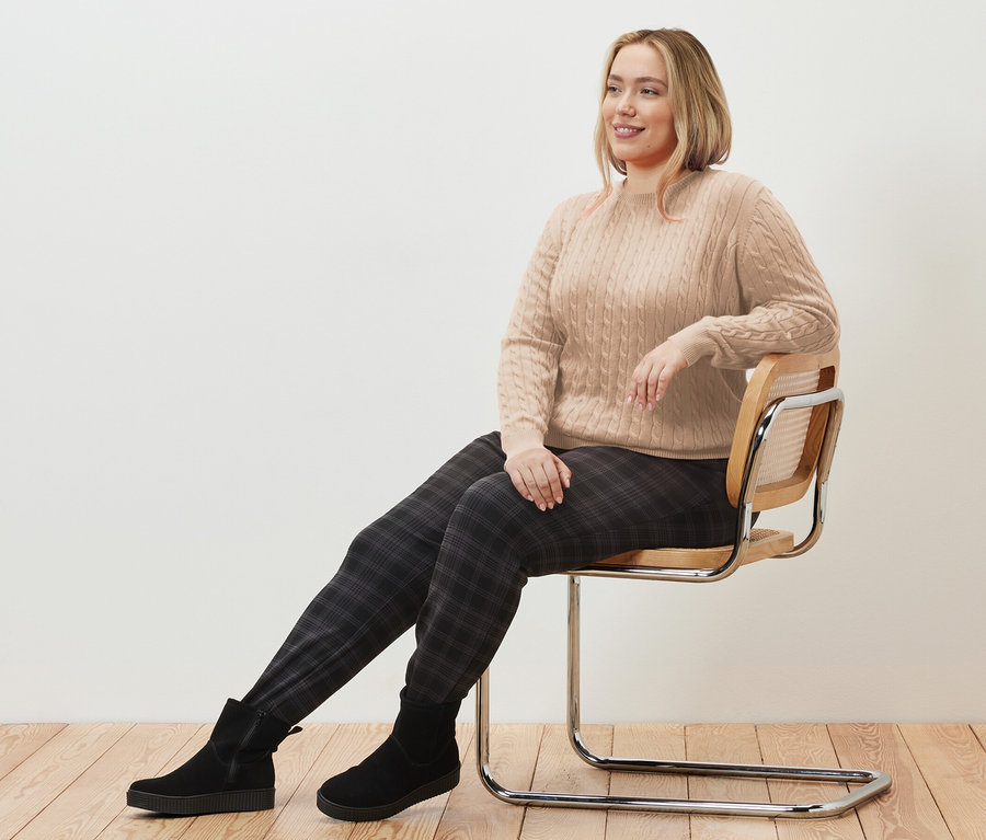 Kobieta siedzi na krześle i ma na sobie kremowy sweter z warkoczowym wzorem i spodnie joggery w kratę.