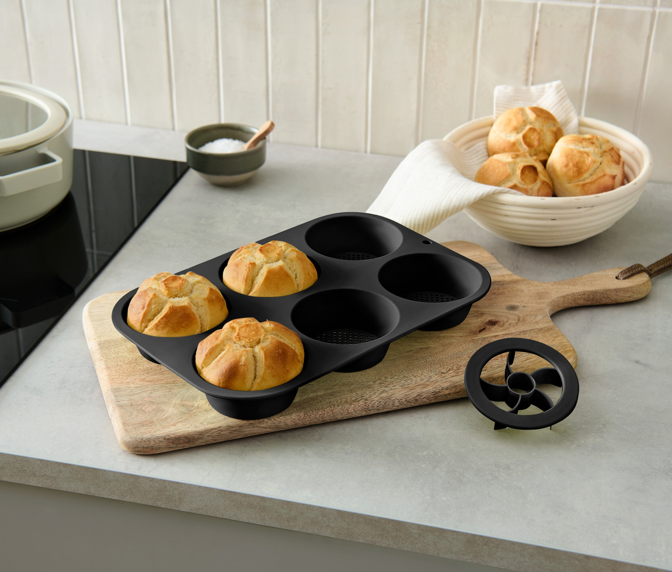 Trzy upieczone bułki w czarnej formie do pieczenia na drewnianej desce obok miski z bułkami.