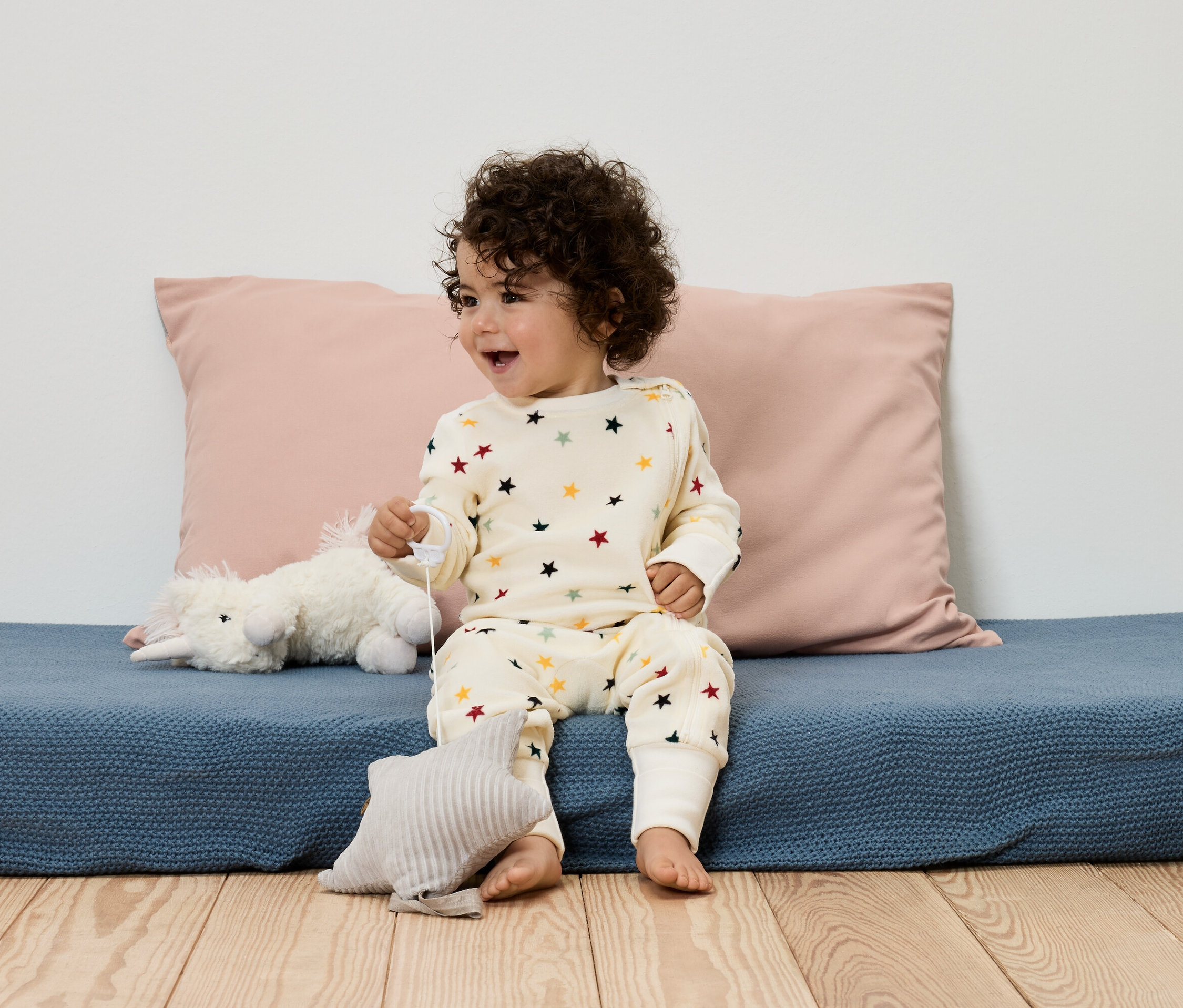 Dziecko siedzi na niebieskiej podkładce w piżamie w gwiazdki, z różową poduszką i zabawkami.